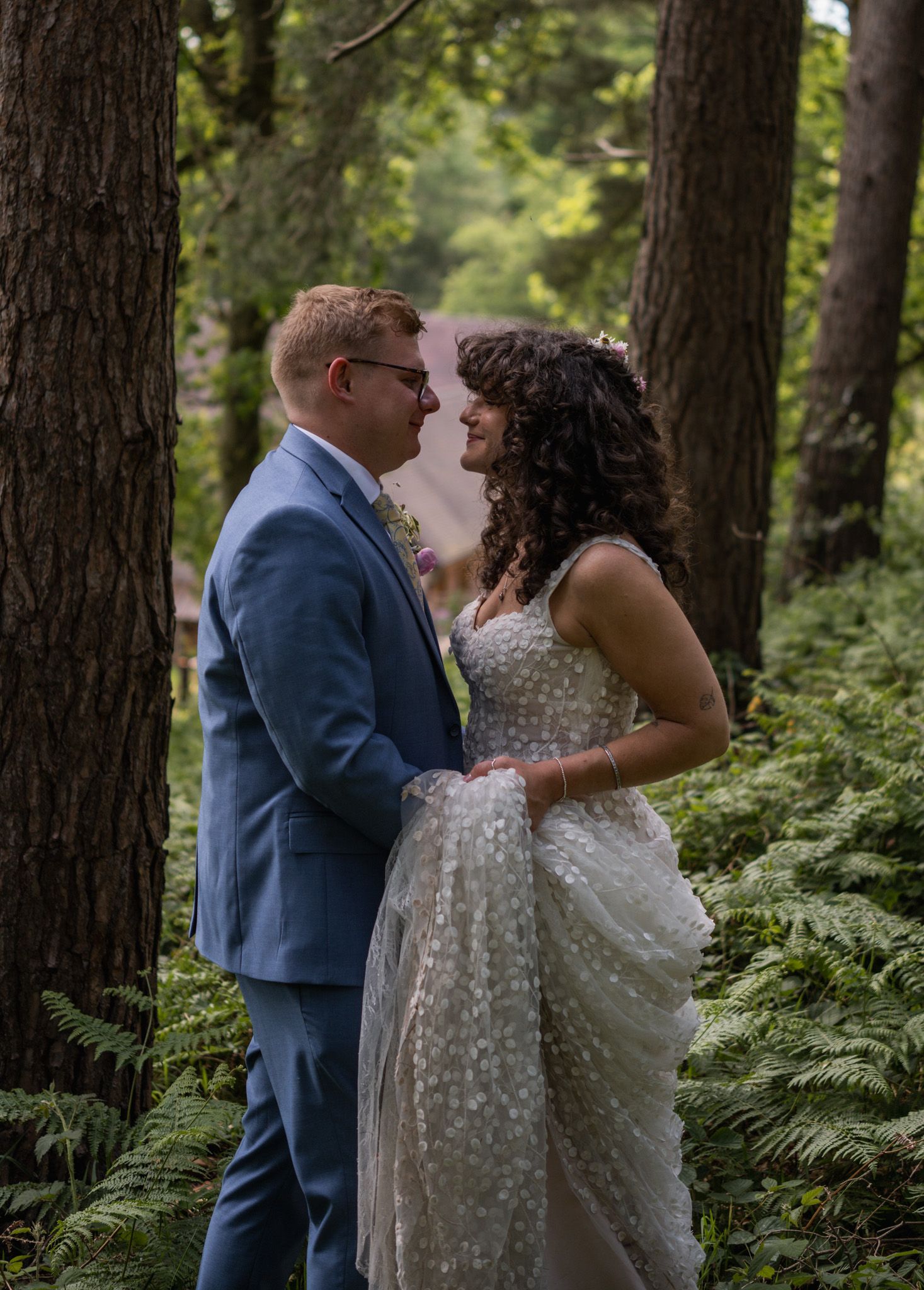 A bride and groom share a romantic moment in a lush forest, surrounded by ferns and tall trees.