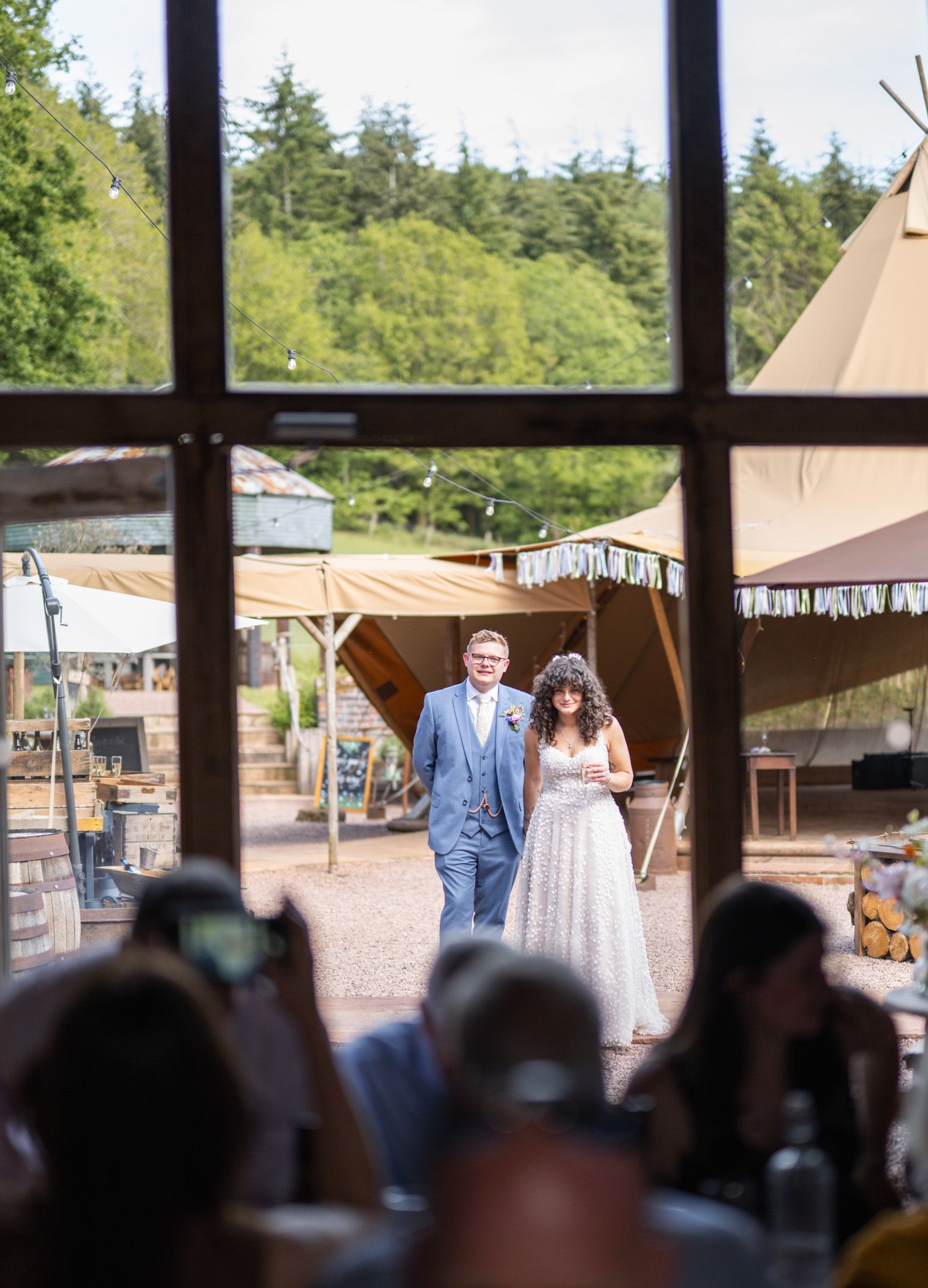Bride and groom walking hand in hand toward a rustic tipi wedding venue surrounded by lush greenery.