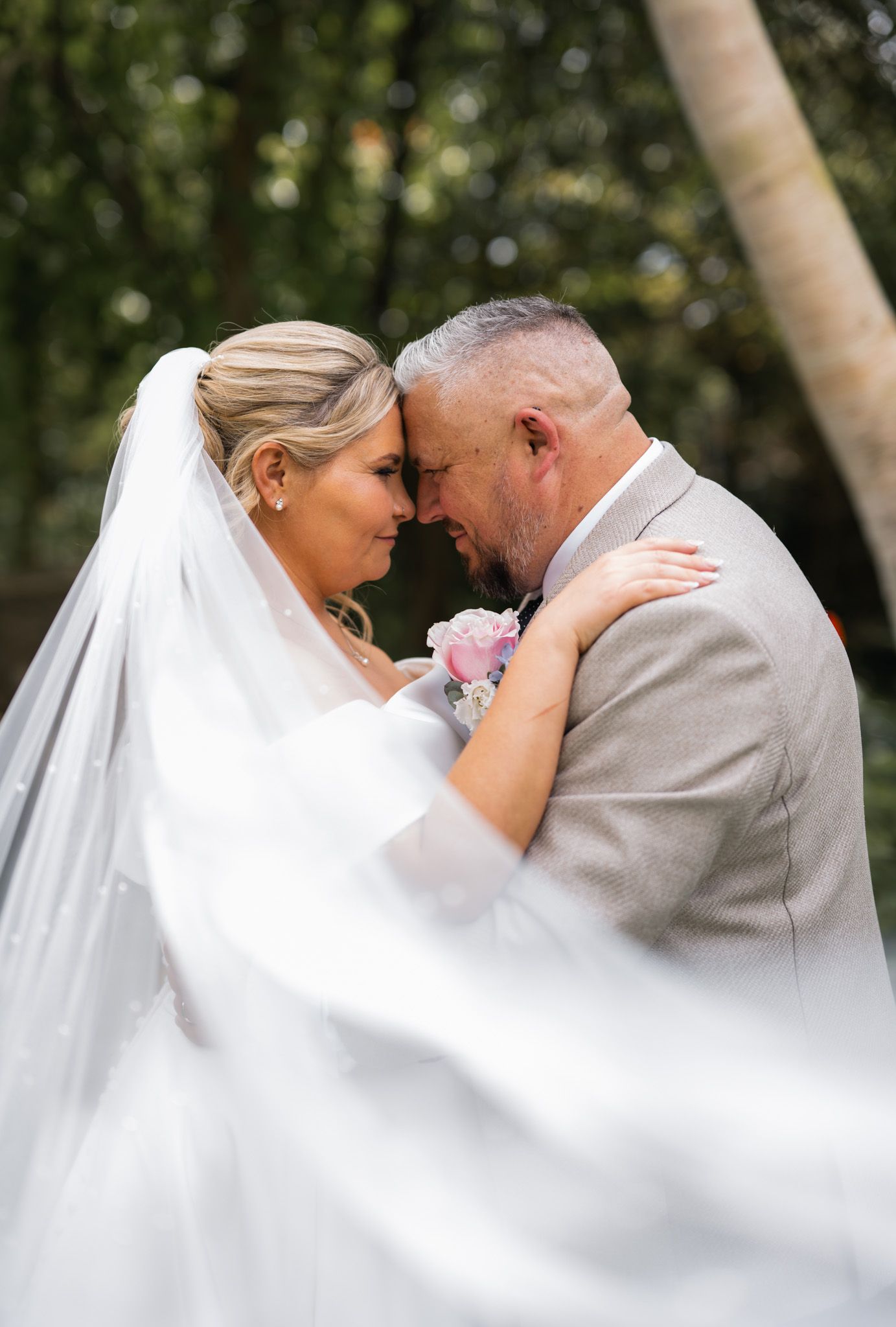 Bride and groom share an intimate forehead touch, her white veil flowing, surrounded by lush greenery at outdoor wedding.
