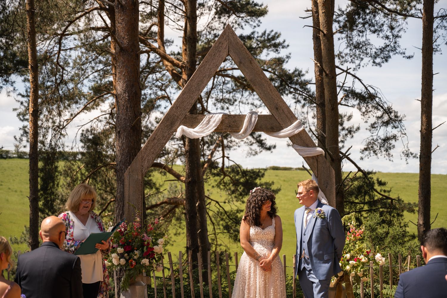 Couple exchanging vows under a wooden triangular arch decorated with flowers at an outdoor wedding ceremony.