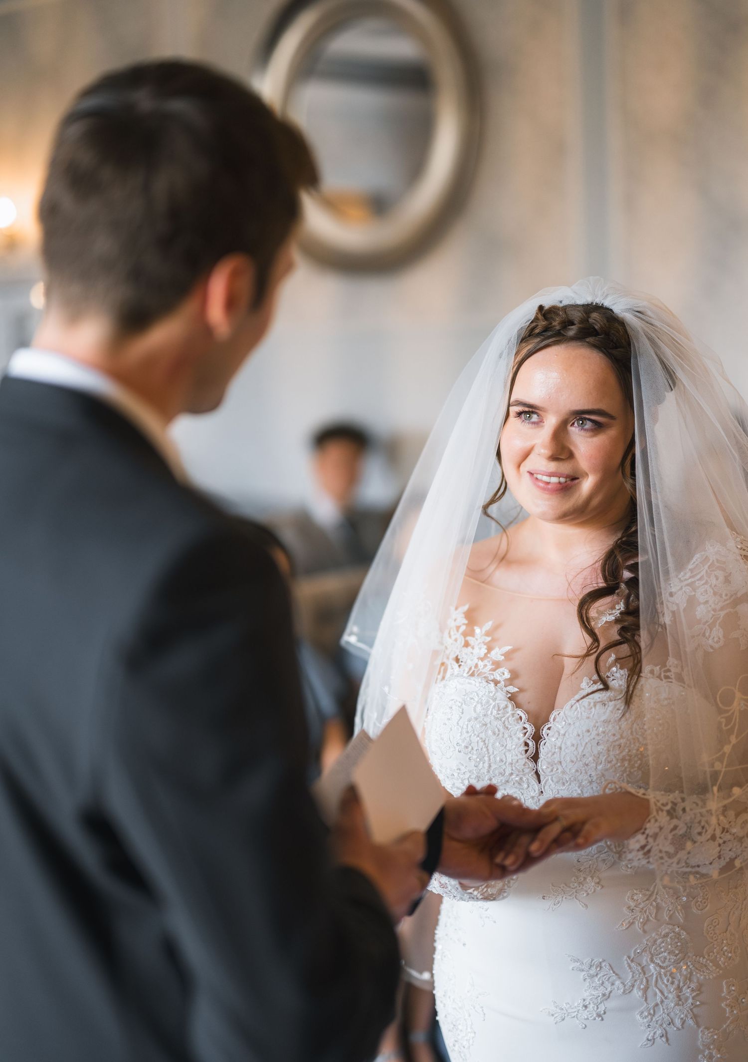 Radiant bride in lace wedding dress and veil smiling at groom during indoor wedding ceremony.