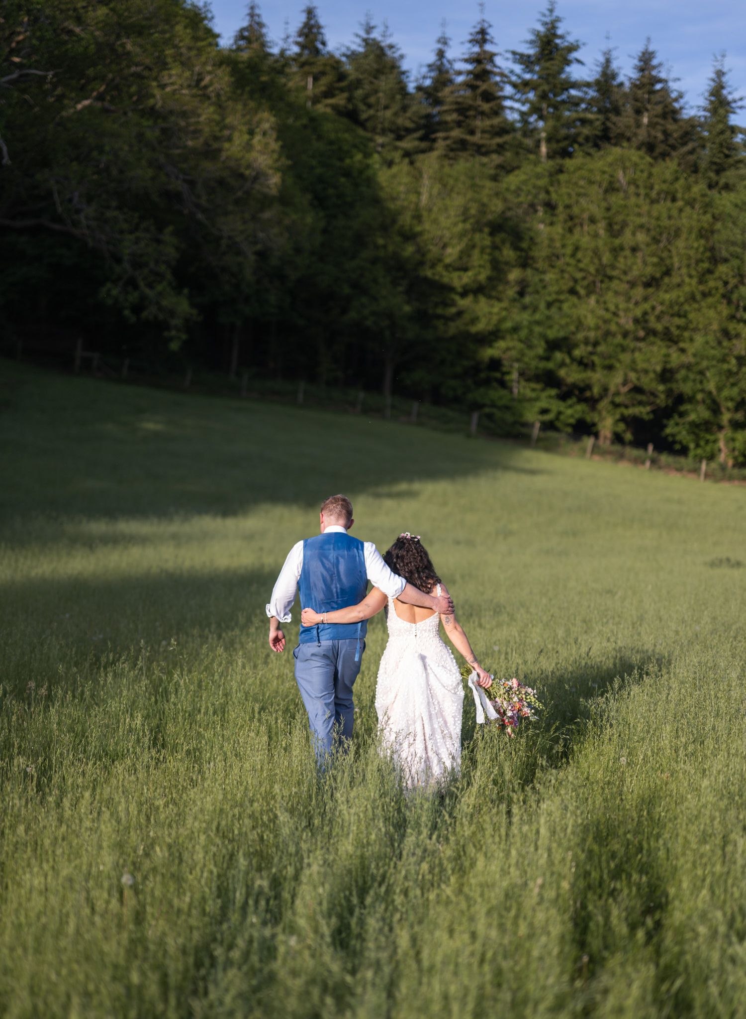 A bride and groom walk hand in hand through a lush green meadow with tall trees in the background on their wedding day.