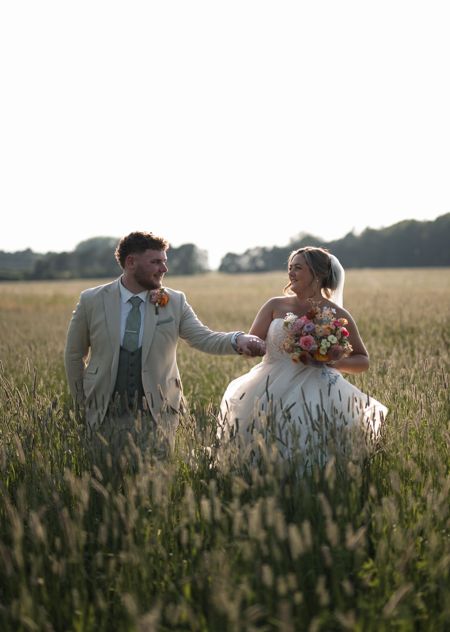 Bride and groom holding hands walking through a golden field at sunset, bride holding a colorful bouquet.