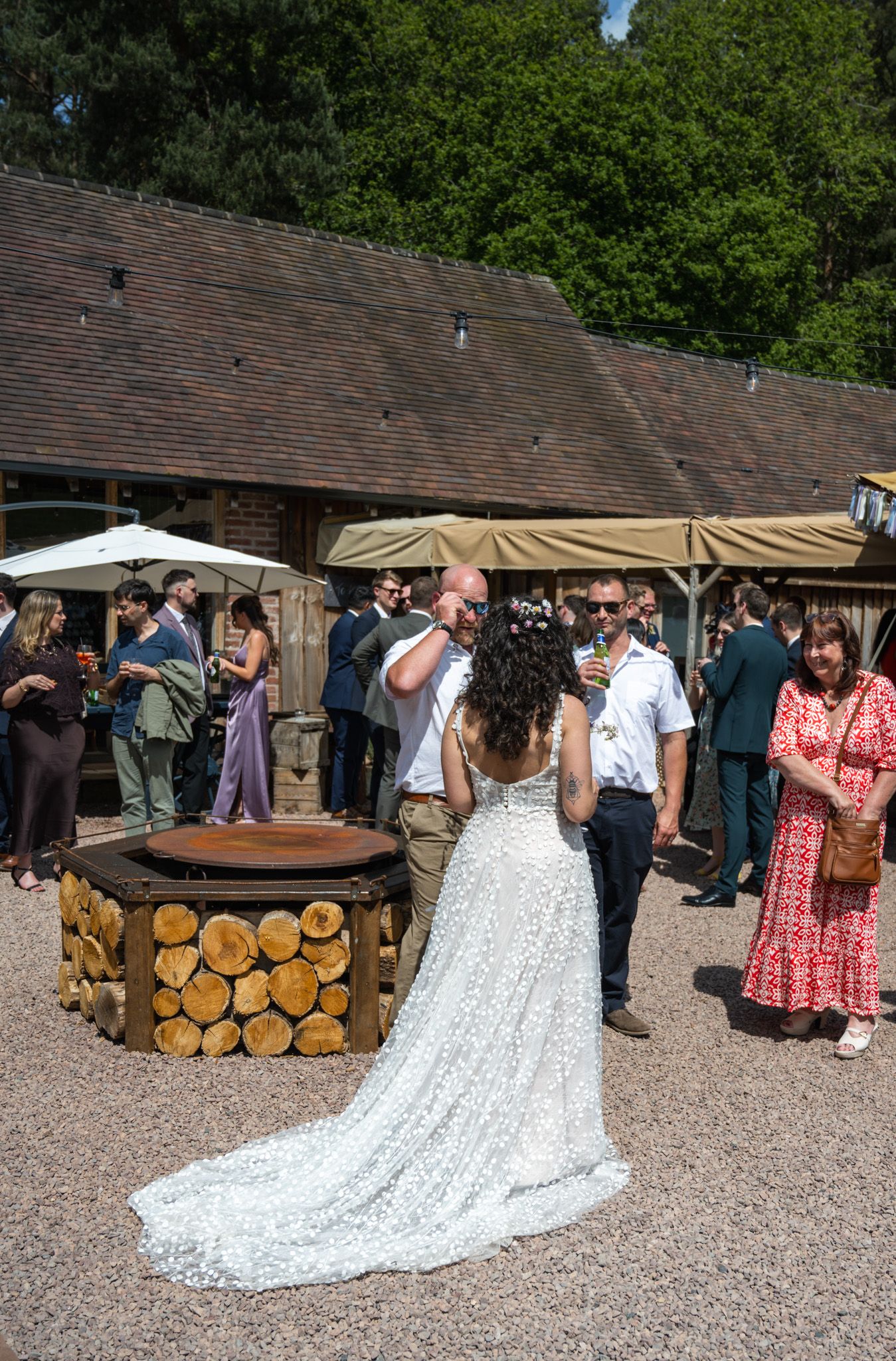 Bride in lace gown with groom at outdoor wedding reception near rustic log fire pit, guests mingling in sunny courtyard.