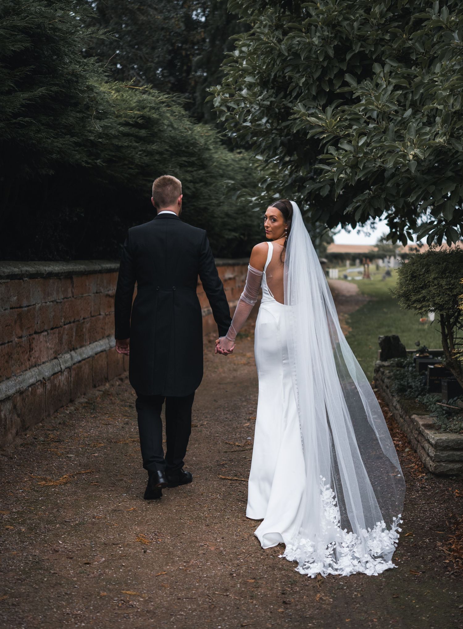 A bride in a white gown with floral veil and groom in black tuxedo walk hand in hand along a garden path.