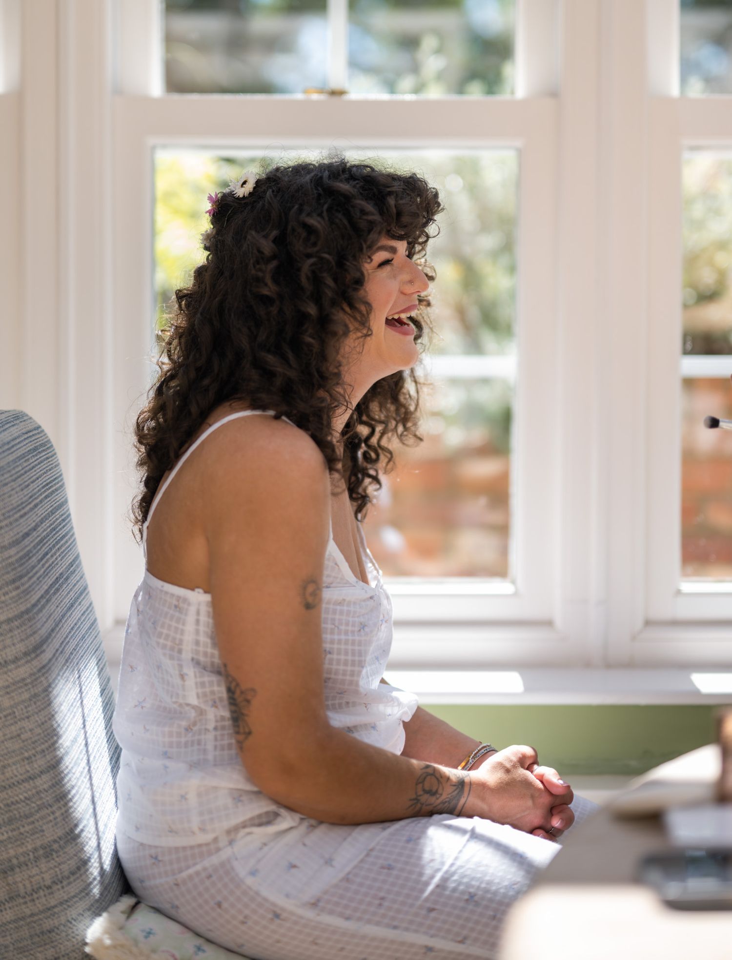 Woman with curly hair laughing joyfully, sitting by a sunlit window in a white dress with tattoos.