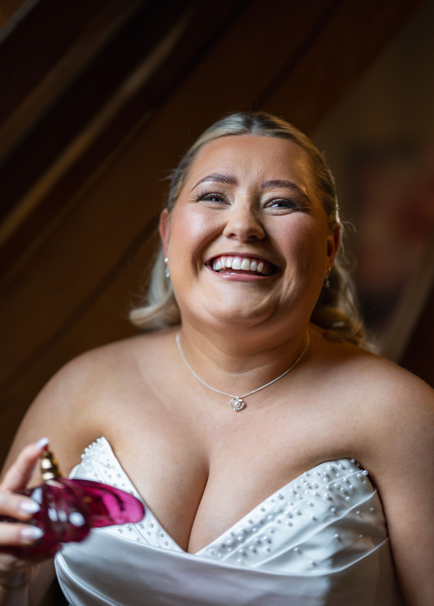 Smiling bride in white strapless gown holding perfume bottle, wearing delicate necklace on her wedding day.