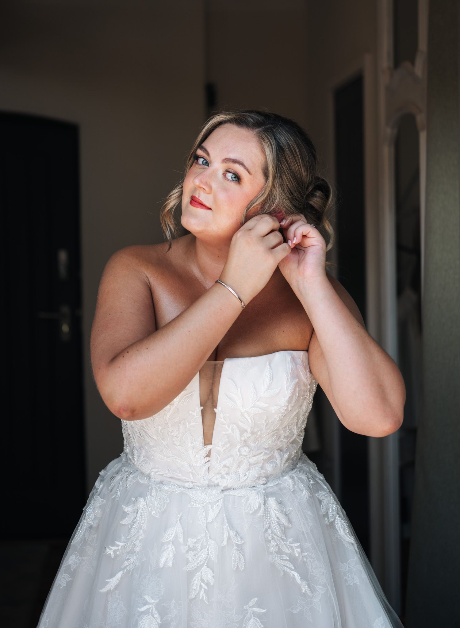 Bride in strapless lace ball gown adjusting earring, with red lips and updo hairstyle, in soft indoor light.
