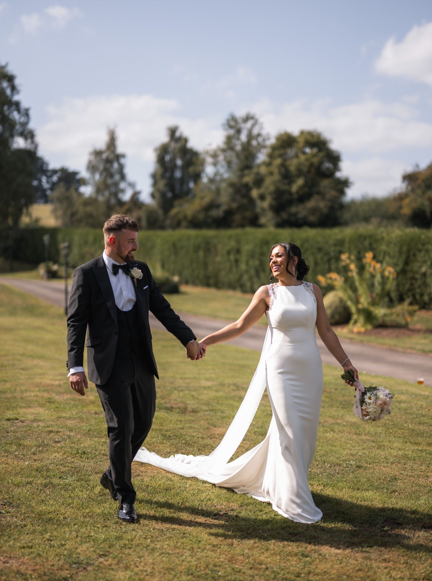 A bride and groom holding hands and walking through lush garden grounds on their sunny wedding day.