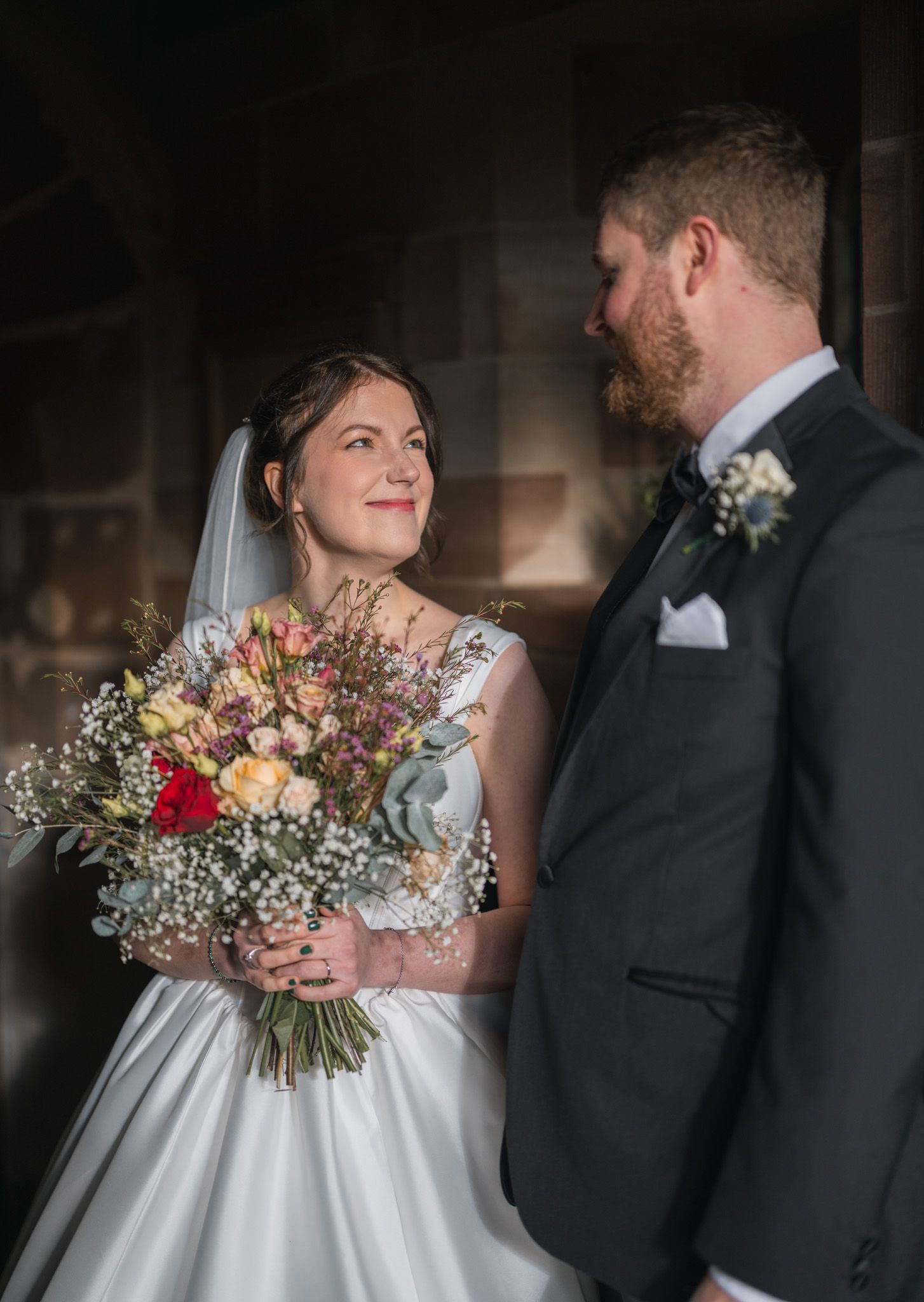 Bride holding colourful bouquet smiles at groom in grey suit during romantic wedding portrait. Shropshire