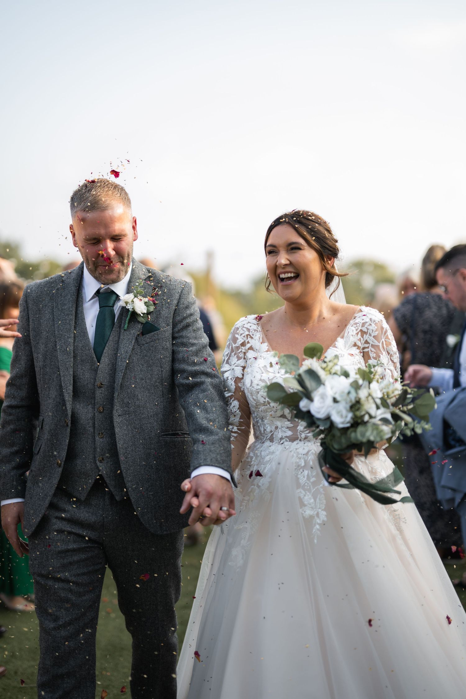 Bride and groom walking through confetti shower, smiling joyfully, bride holding white bouquet in elegant lace wedding dress.