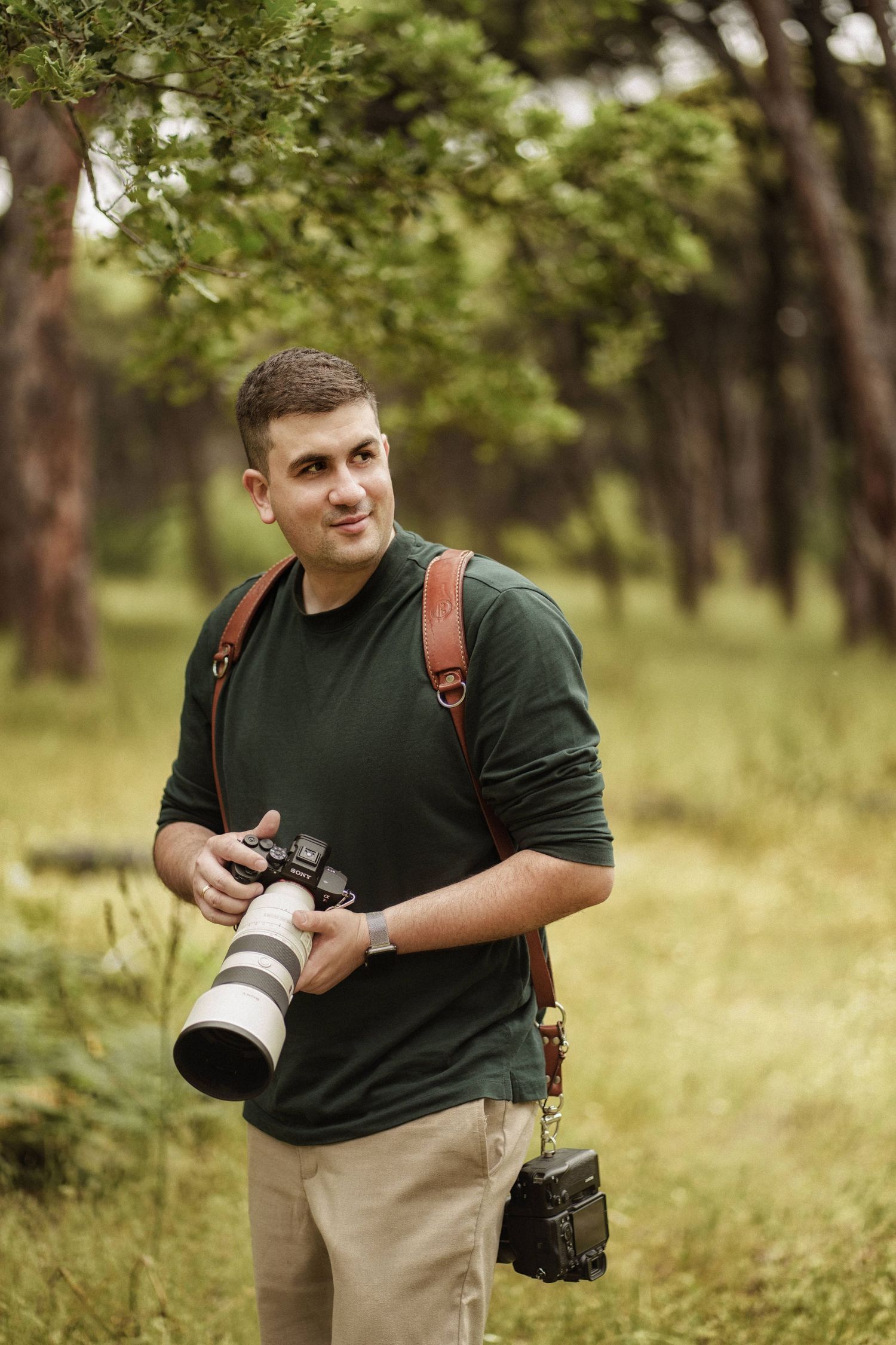 Professional photographer wearing green shirt carries camera equipment while walking through scenic wooded area.