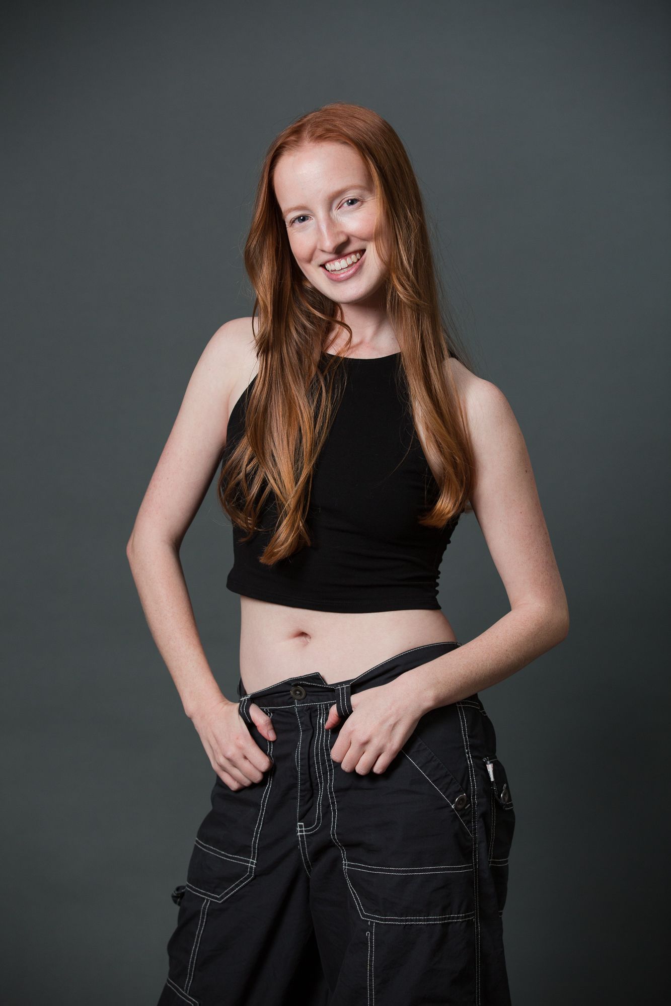 Smiling young woman with long red hair wearing a black crop top and dark cargo pants against a gray background.