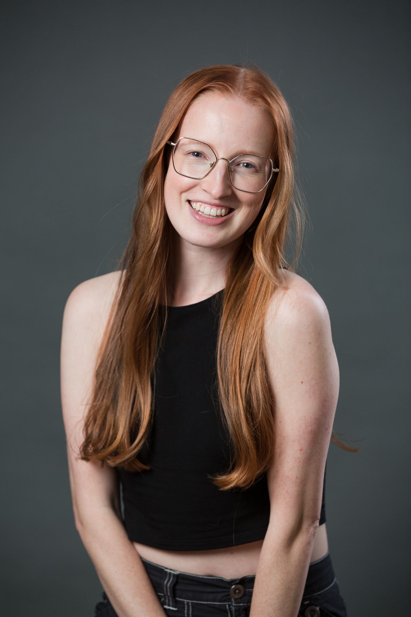 Smiling young woman with long red hair and glasses wearing a black crop top against a gray background.