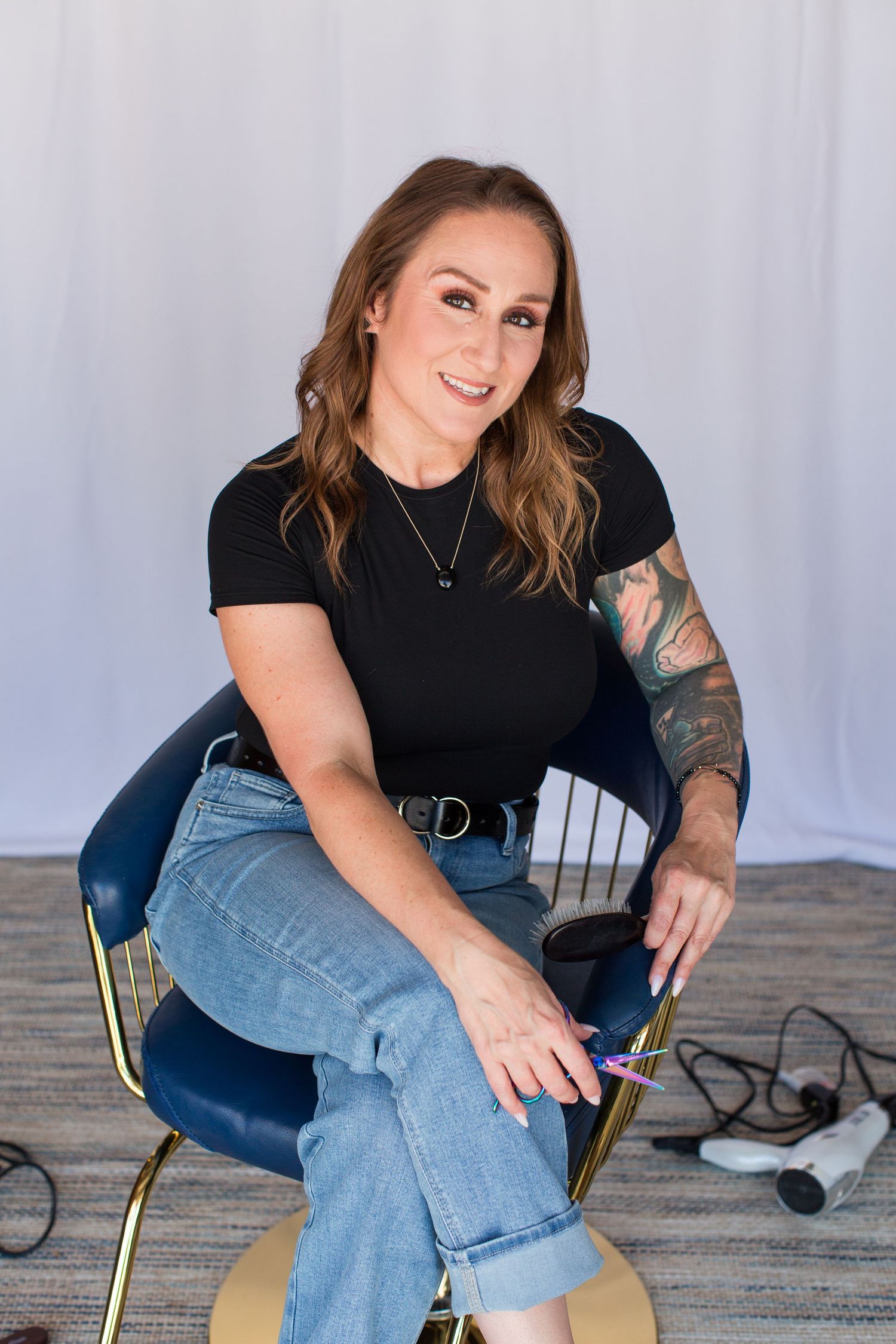 Person in black shirt and jeans sitting casually on a chair against white backdrop showing tattoos.