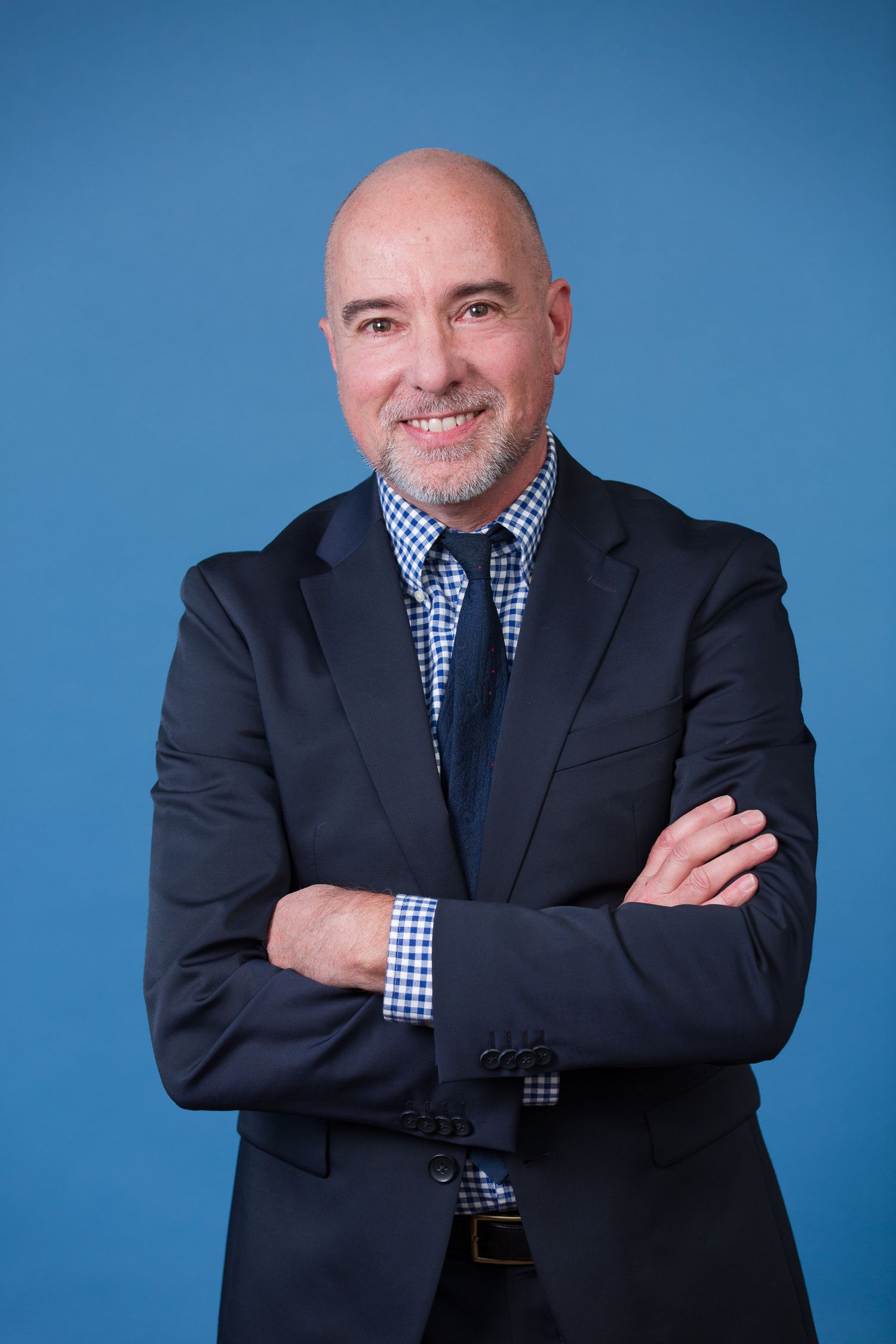 Professional studio portrait against blue backdrop showing confident pose in business attire with checkered shirt.