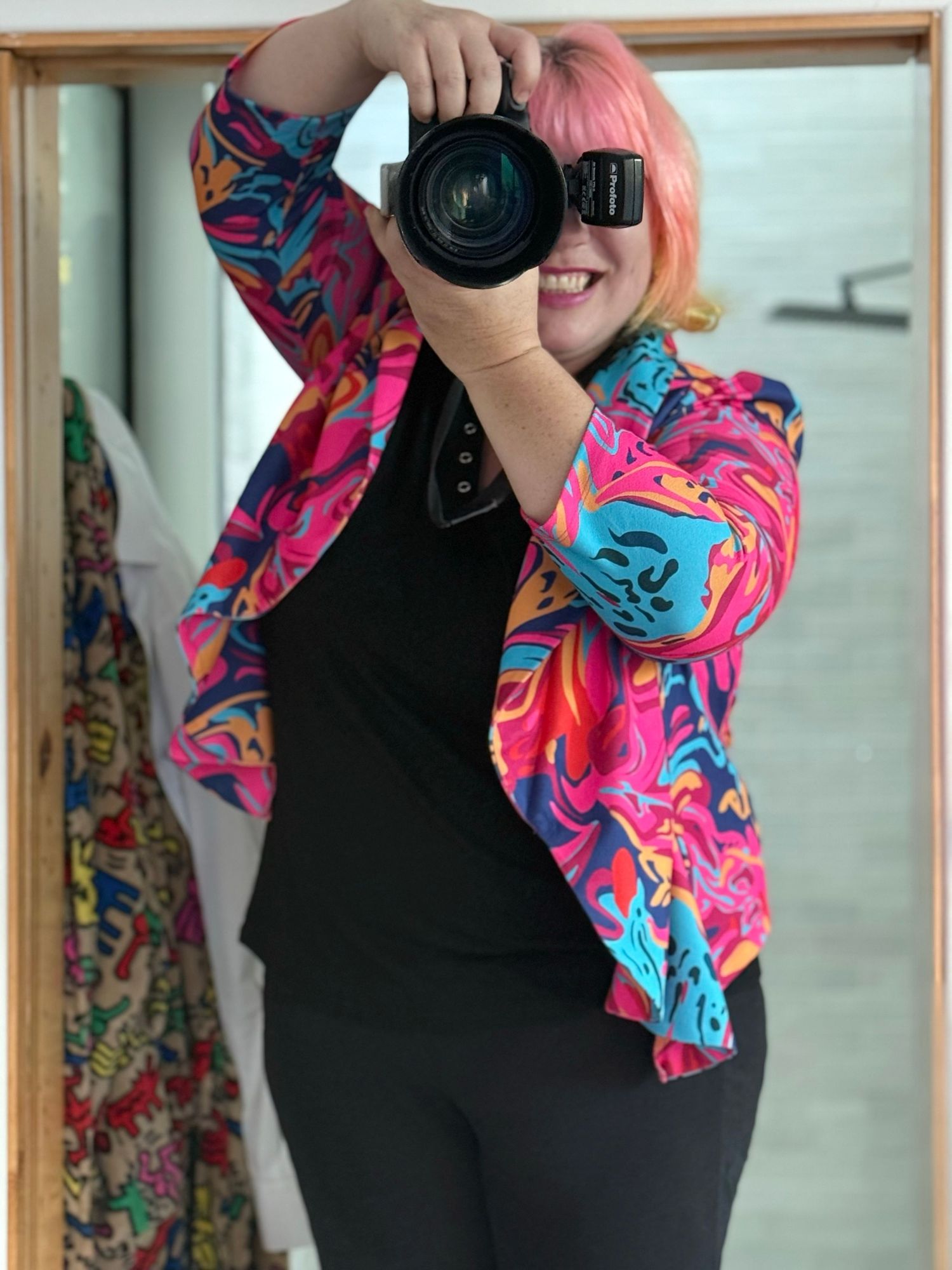 Photographer in black outfit with vibrant pink and blue patterned scarf holds camera in artistic poses.