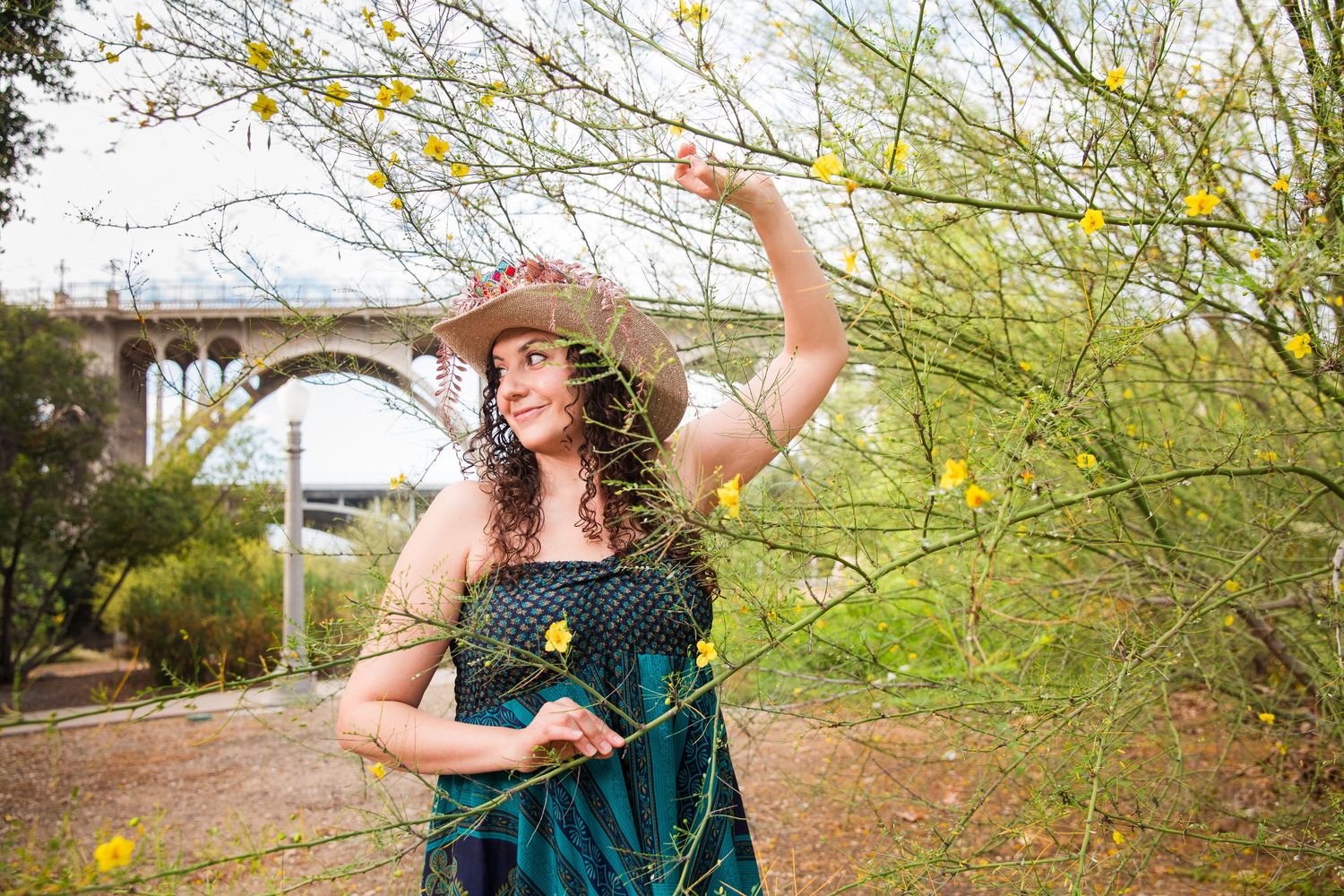Smiling woman with curly hair and a straw hat raises one arm among yellow wildflowers, with a stone arch bridge in the background.