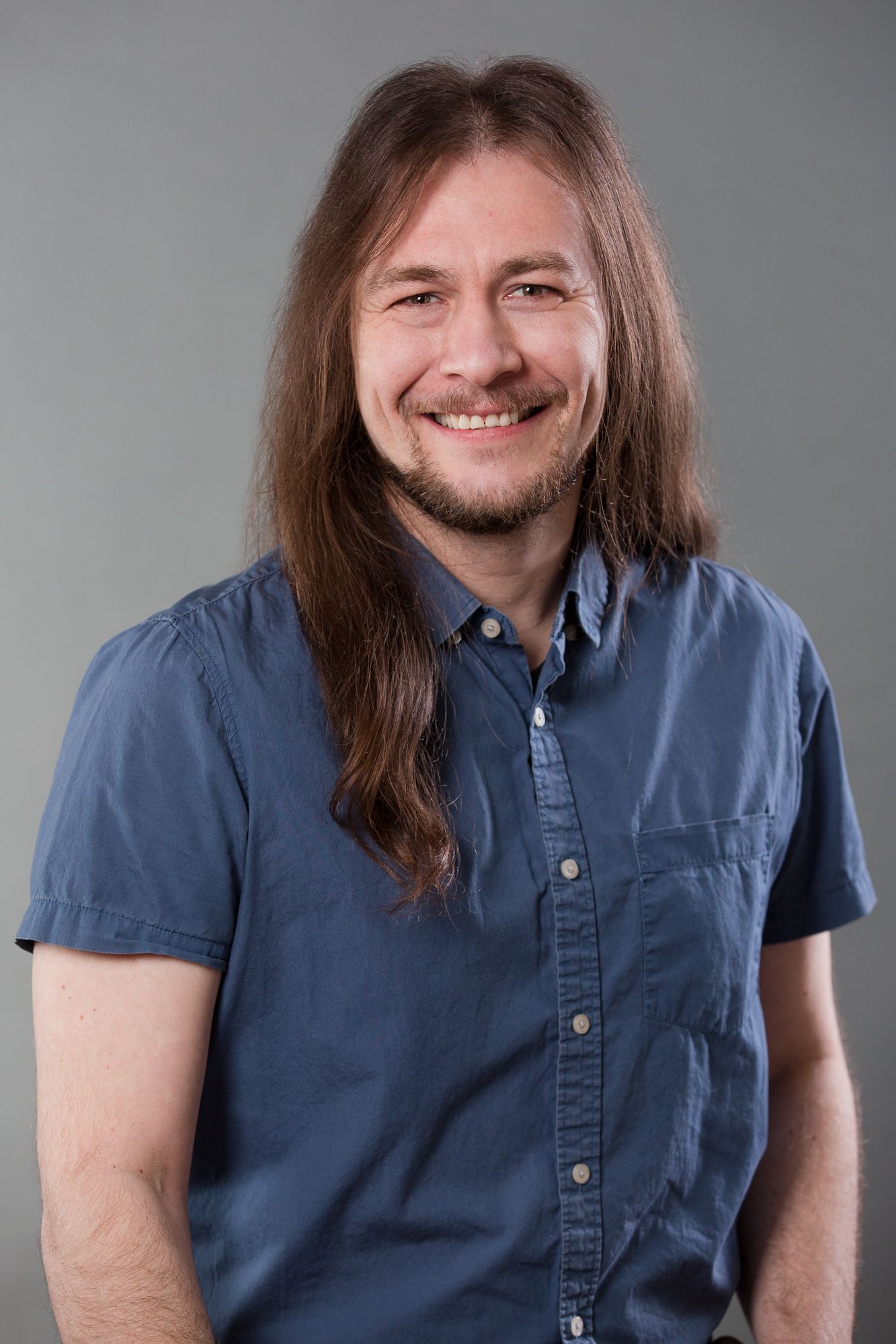 Smiling man with long brown hair and goatee wearing a blue short-sleeve button-up shirt against a gray background.