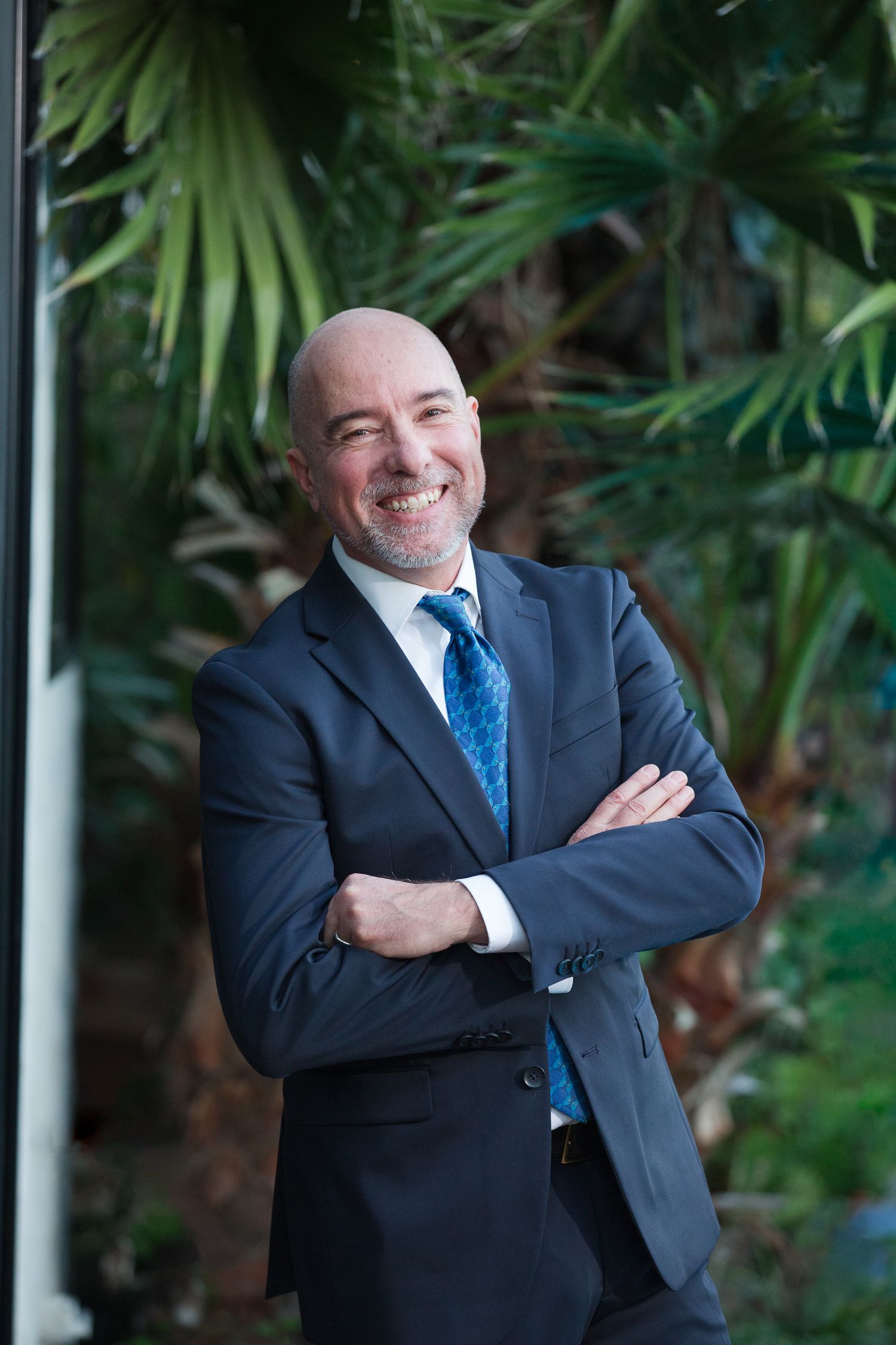 Professional in a dark suit and blue tie poses with crossed arms against a green foliage background.