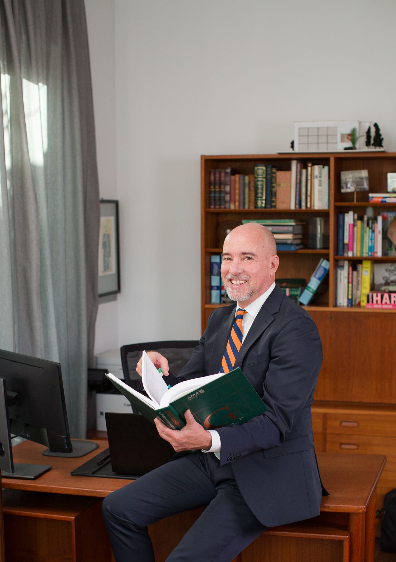Professional in dark suit sits at wooden desk in office with bookshelves and reads through documents.