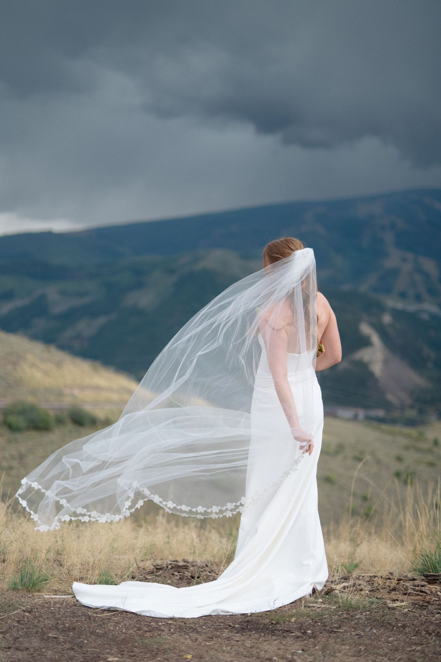 A bride in a flowing white dress and veil stands on a mountaintop with dramatic storm clouds in the background.