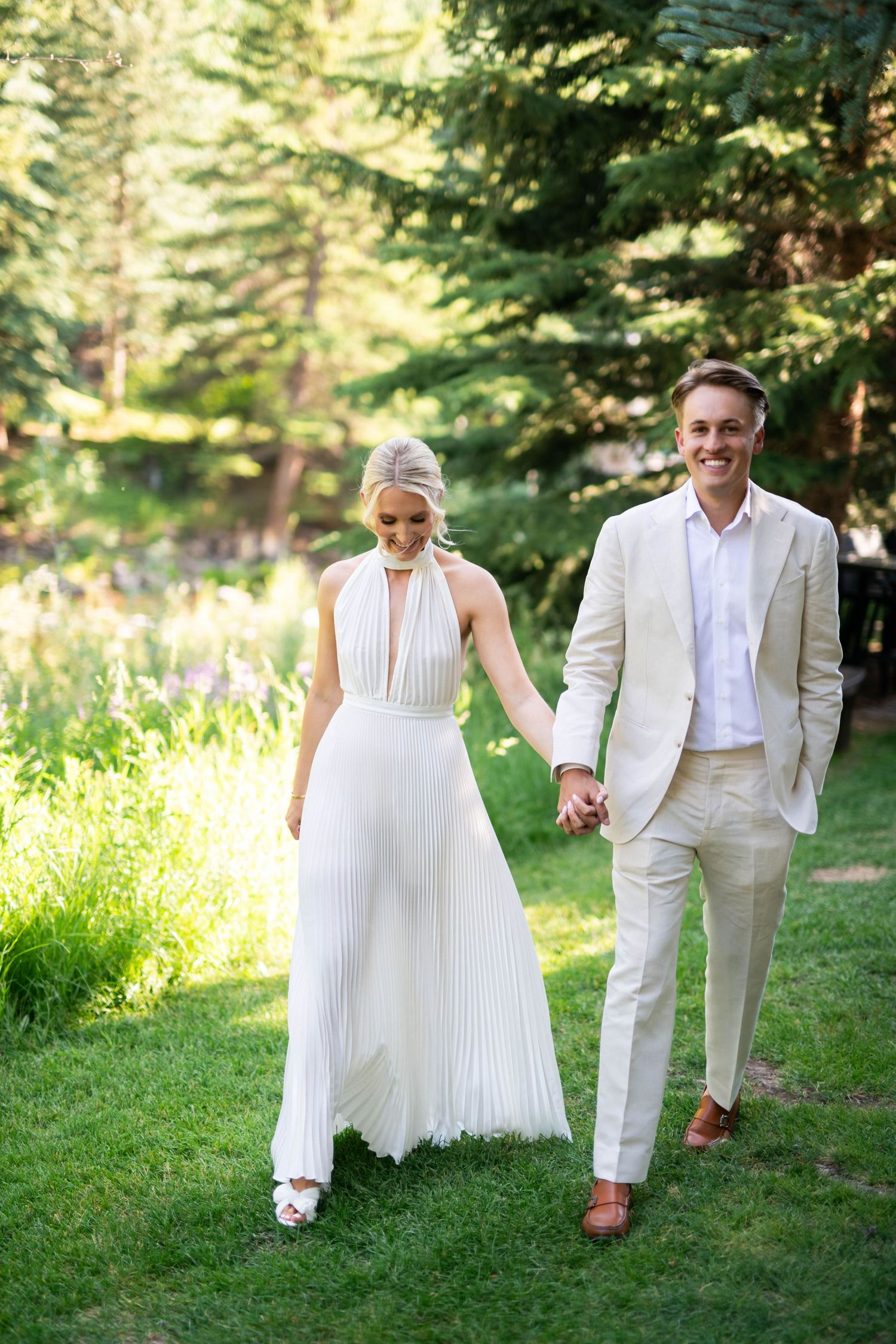 A couple in matching white outfits playfully poses in a sunlit forest setting.
