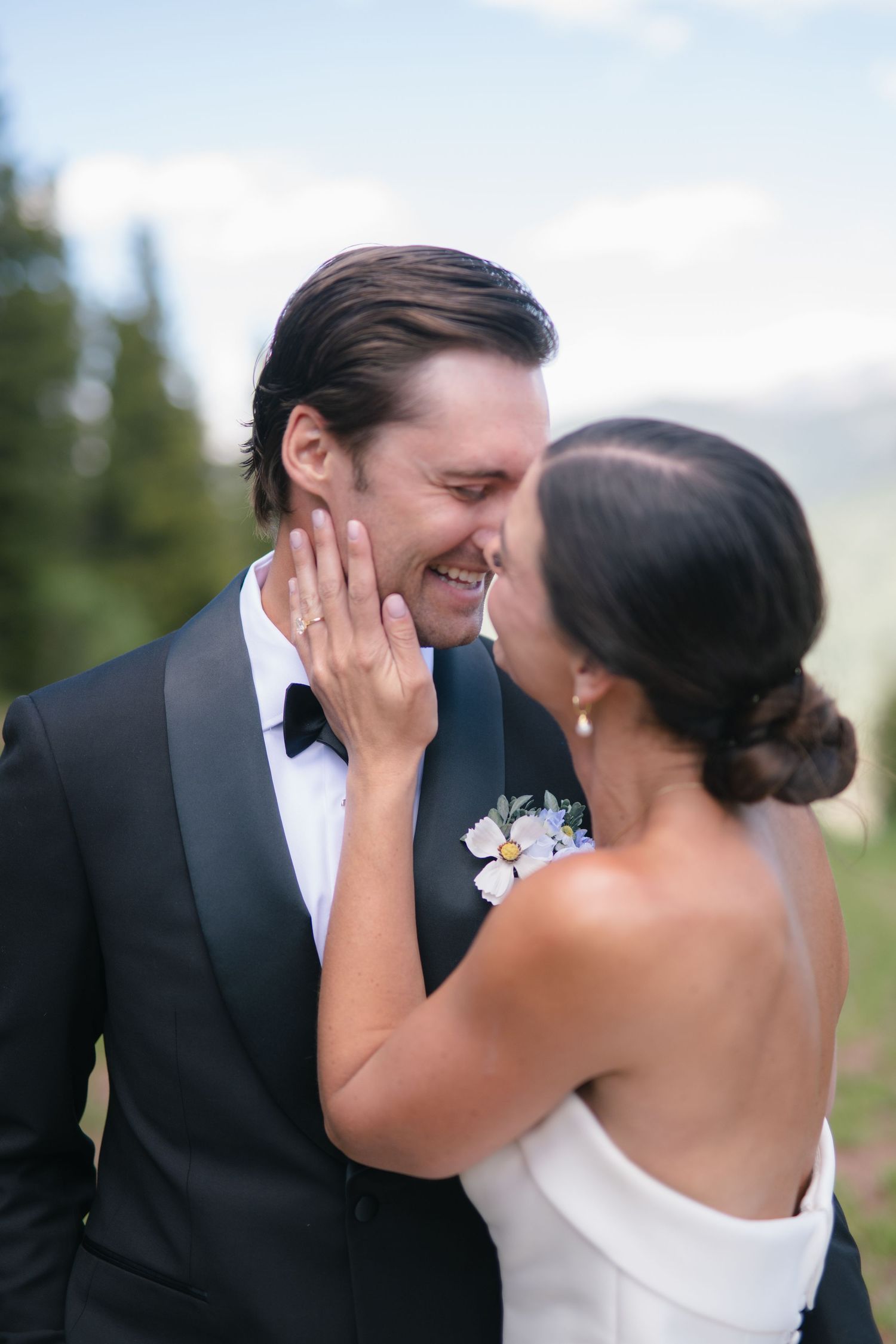 A joyful bride and groom share an intimate moment, smiling close together, with the groom in a black tuxedo outdoors.
