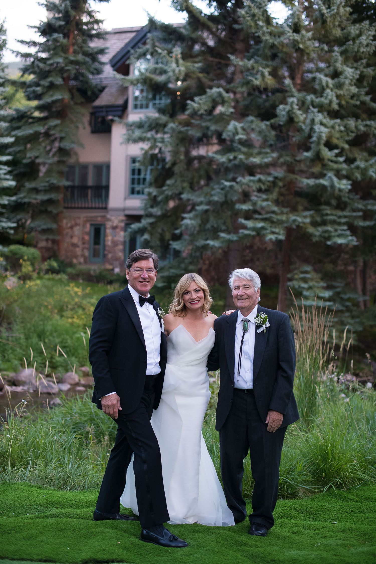 Bride in white gown poses with two men in tuxedos outdoors at elegant wedding venue surrounded by pine trees.