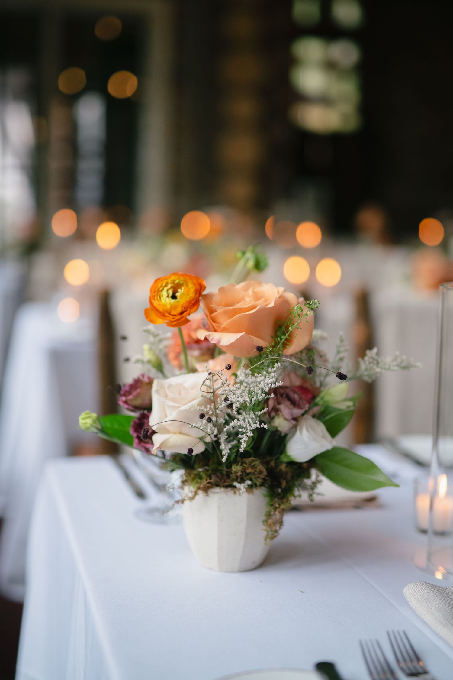 Elegant floral centerpieces with peach ranunculus and white roses on white tablecloths at a wedding reception.