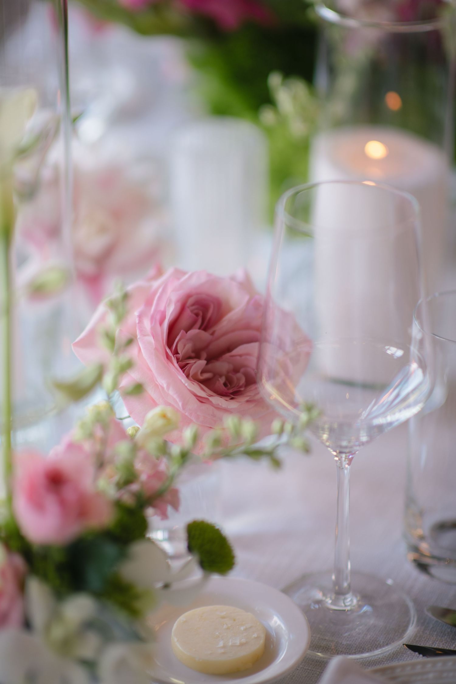 Elegant wedding table setting with pink roses, crystal wine glass, white candle, and a small macaron on a white plate.