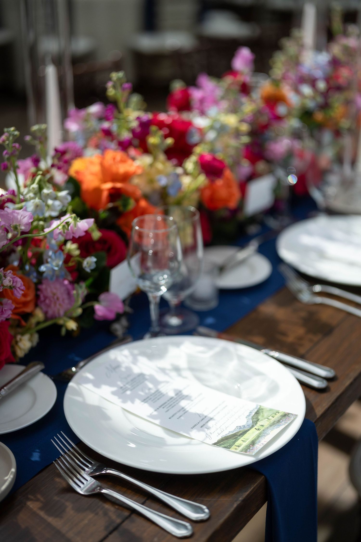 Elegant wedding table setting with navy runner, white plates, silverware, and vibrant floral centerpieces in pink, orange, and red.