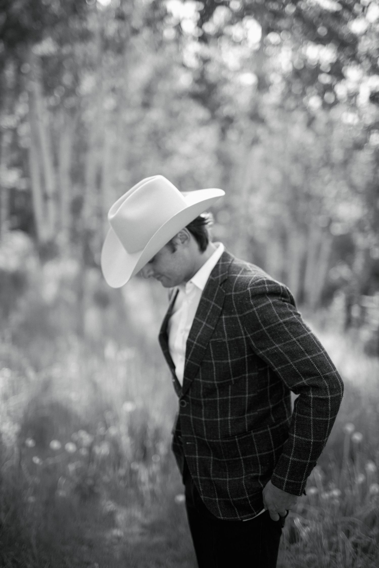 A person in a cowboy hat and plaid blazer stands in profile against a blurred nature background.