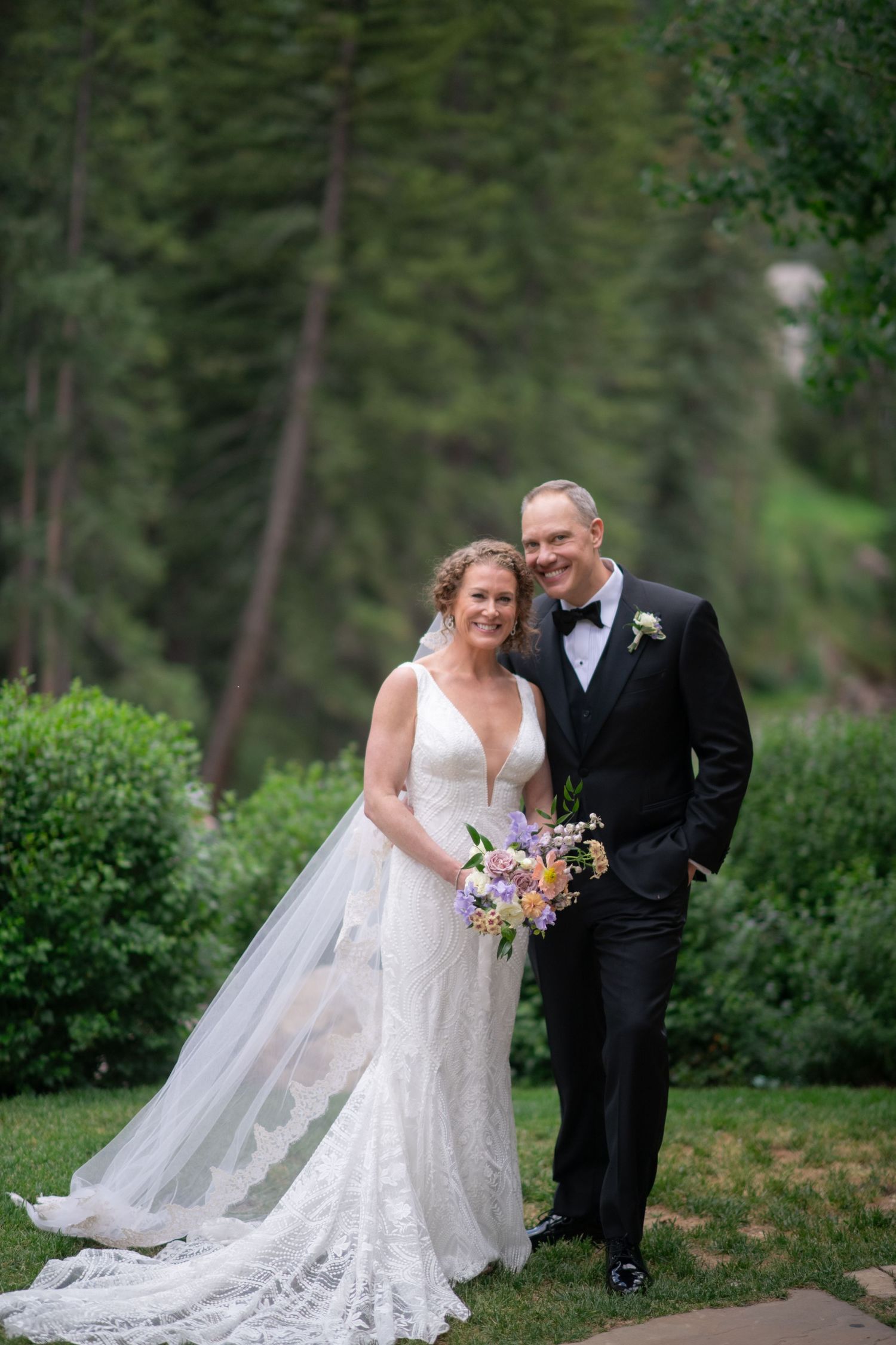 Newlywed couple poses in forest setting with bride wearing lace dress and flowing veil.