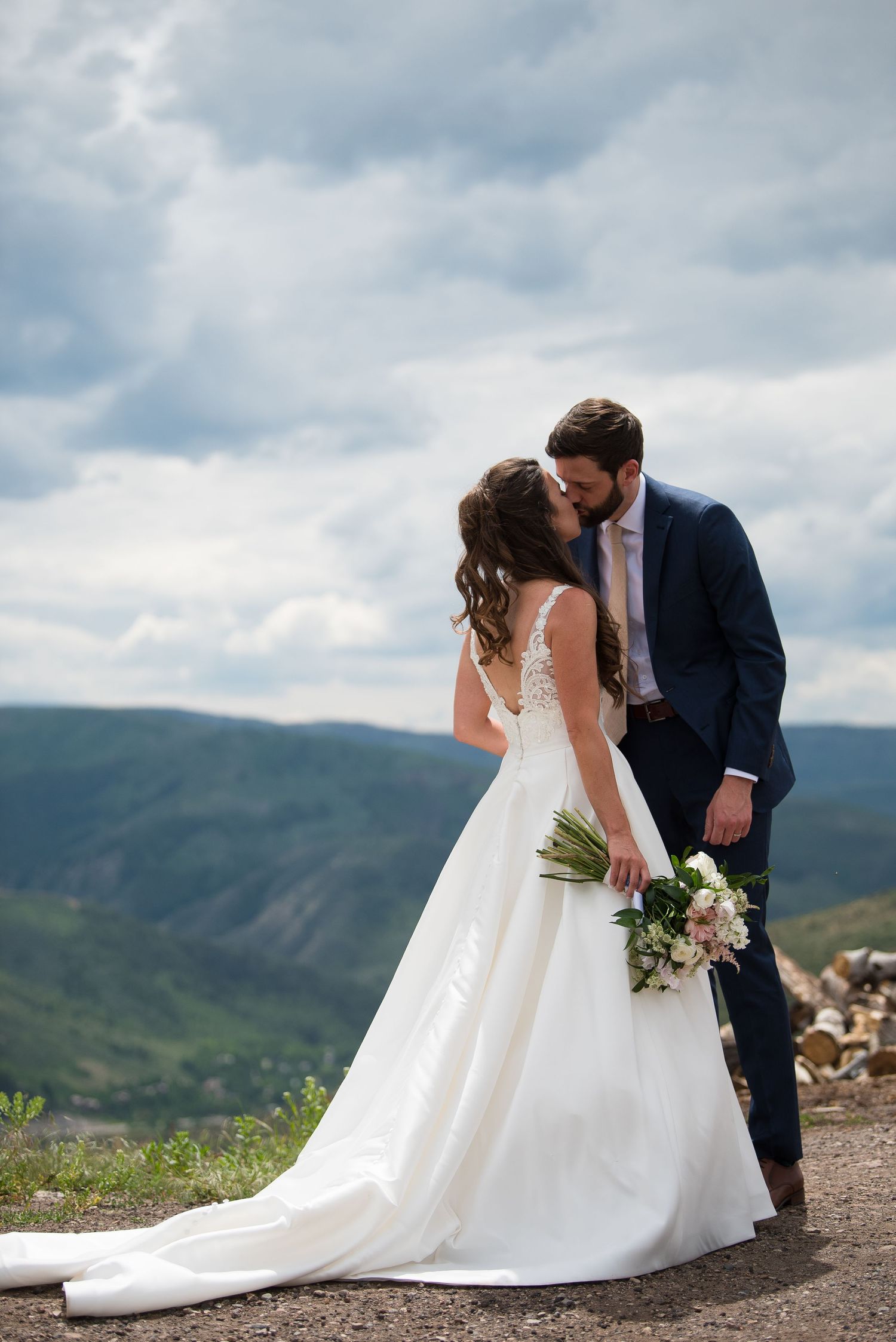 Bride and groom share a kiss on a mountain overlook, she holds a bouquet in her white gown as he wears a navy suit.