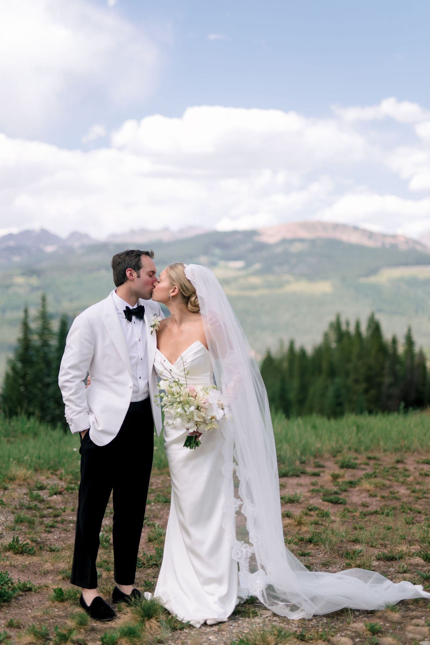 Bride and groom share a kiss on a mountain overlook, with evergreen trees and Rocky Mountain peaks in the background.