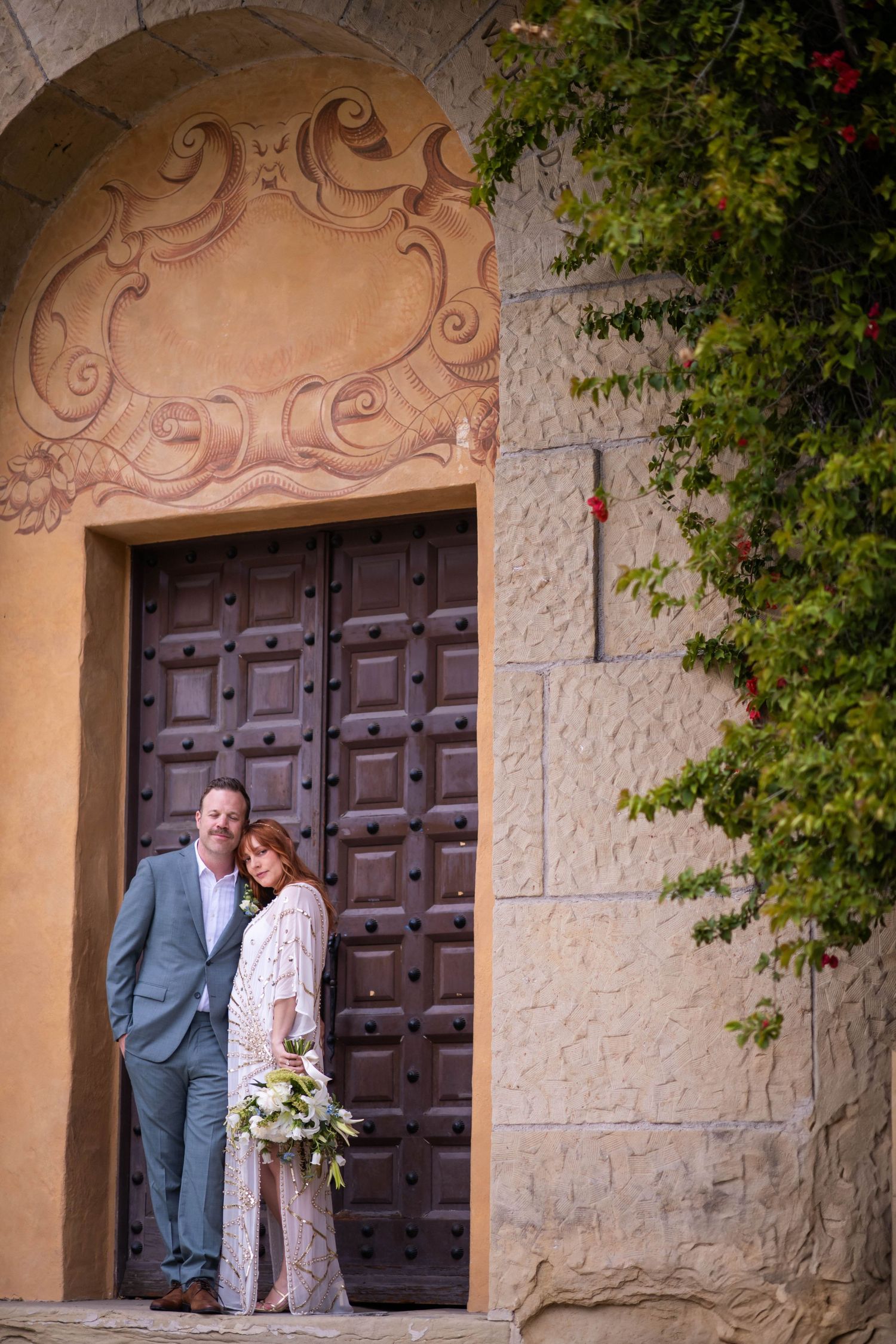 Wedding couple shares intimate moment against stone archway with ornate wooden doors.