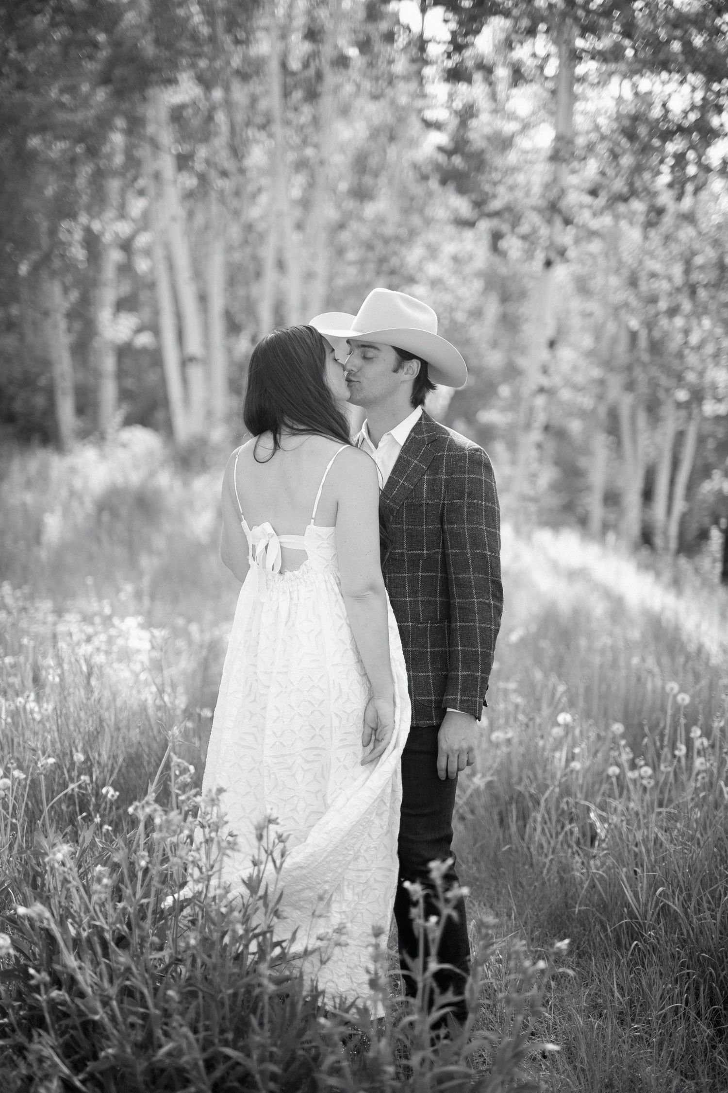 Black and white photo of a couple kissing in a meadow, bride in white dress, groom in plaid suit and cowboy hat.