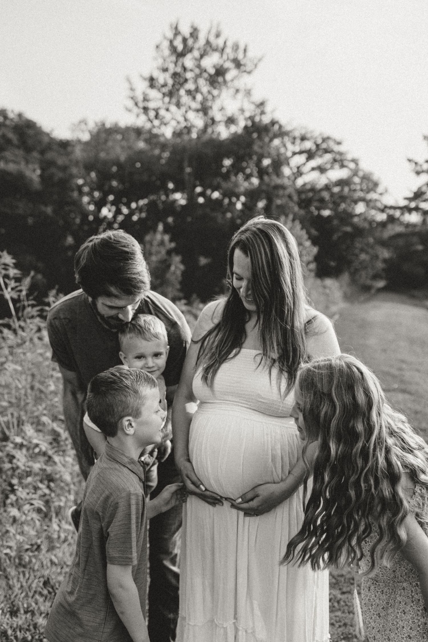 A family shares tender moments outdoors in a field, touching a pregnant belly in a series of black and white photos.