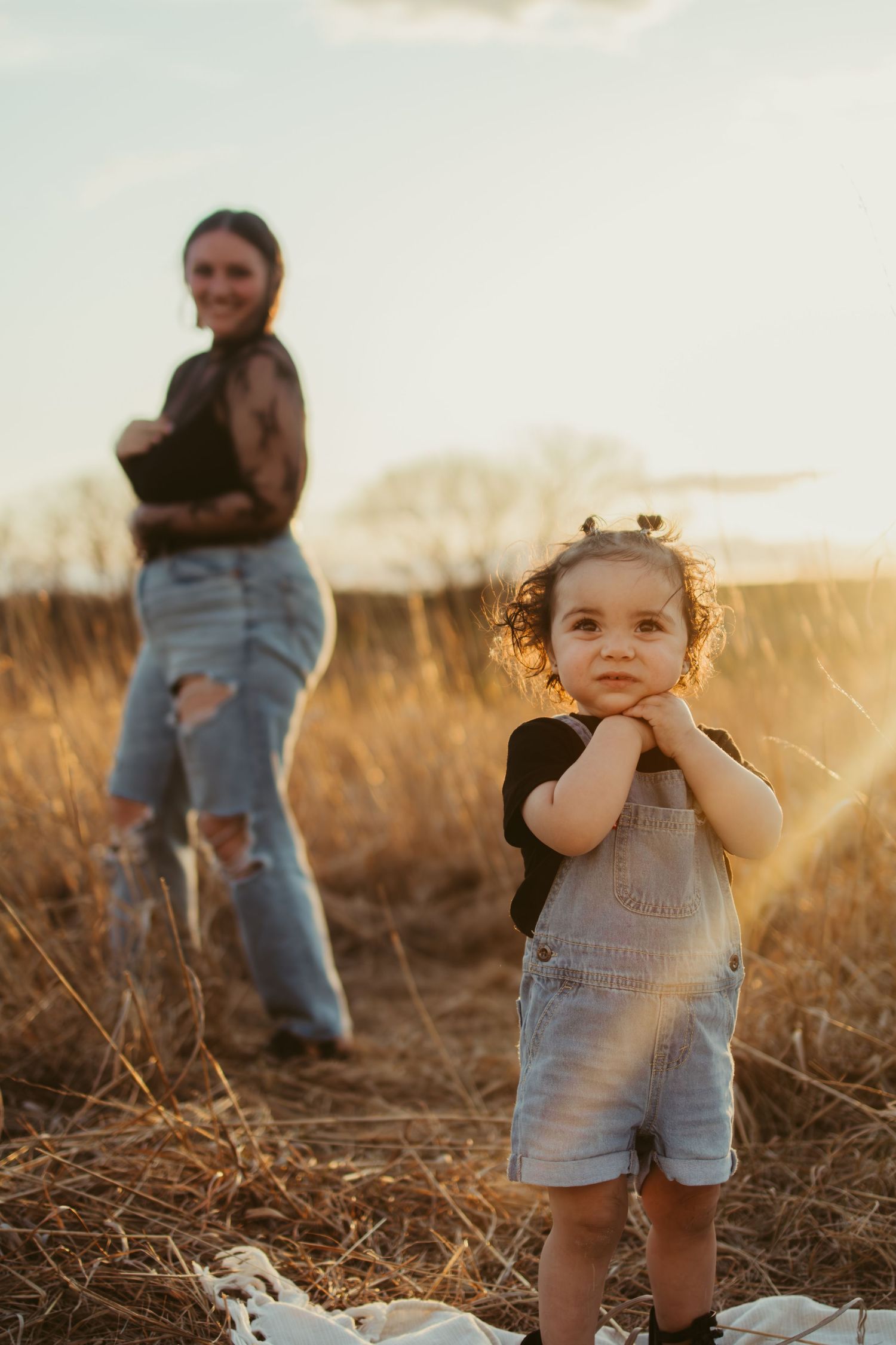 Young child in denim overalls poses playfully at sunset in a field of tall grass.