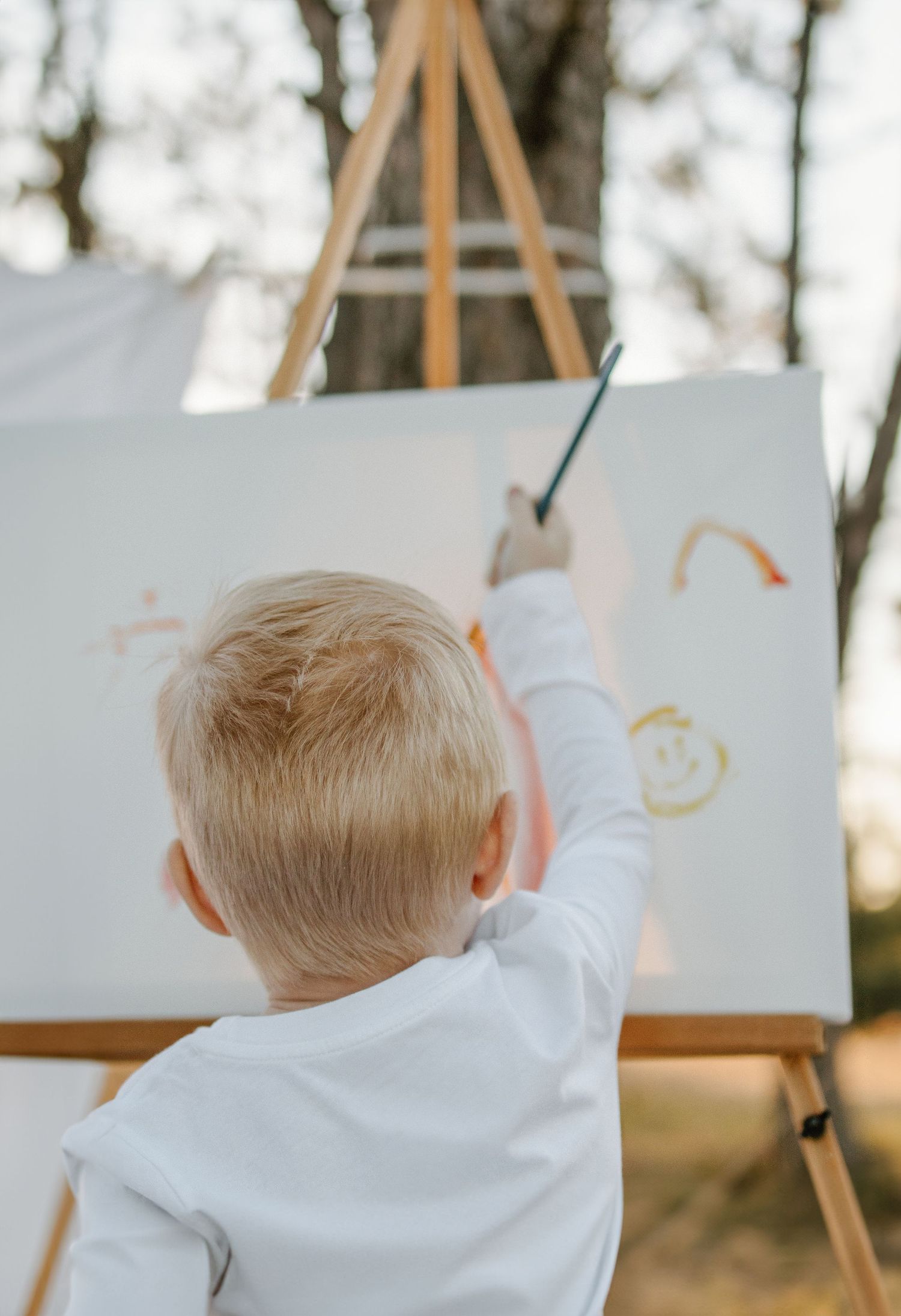 Back view of small child in white shirt drawing on easel outdoors.