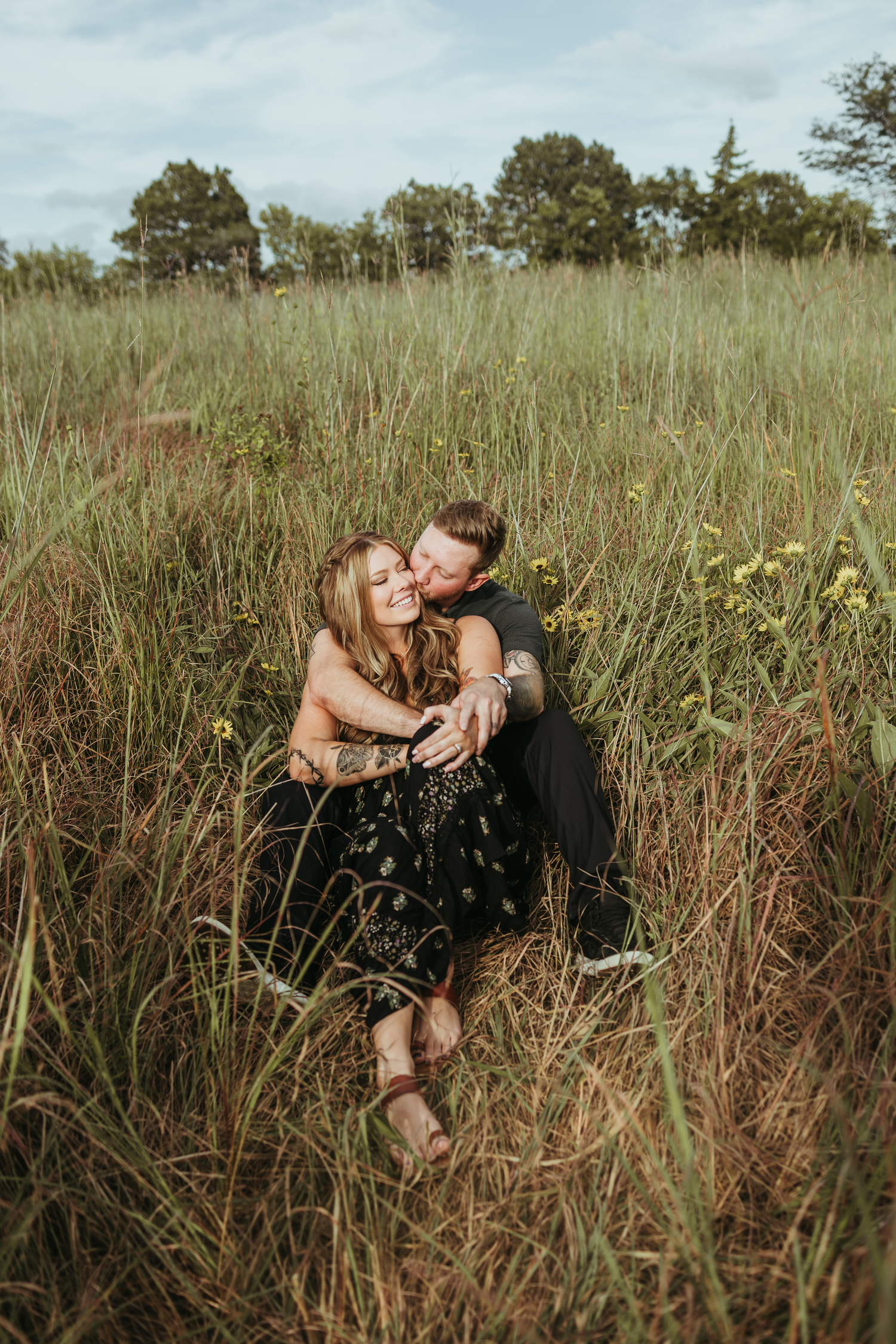 A couple shares intimate moments while sitting together in a tall grass field with wildflowers on a cloudy day.