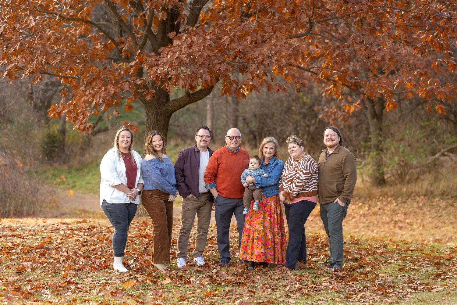 A group of people sit together on a fallen tree trunk surrounded by vibrant autumn foliage in a forest setting.