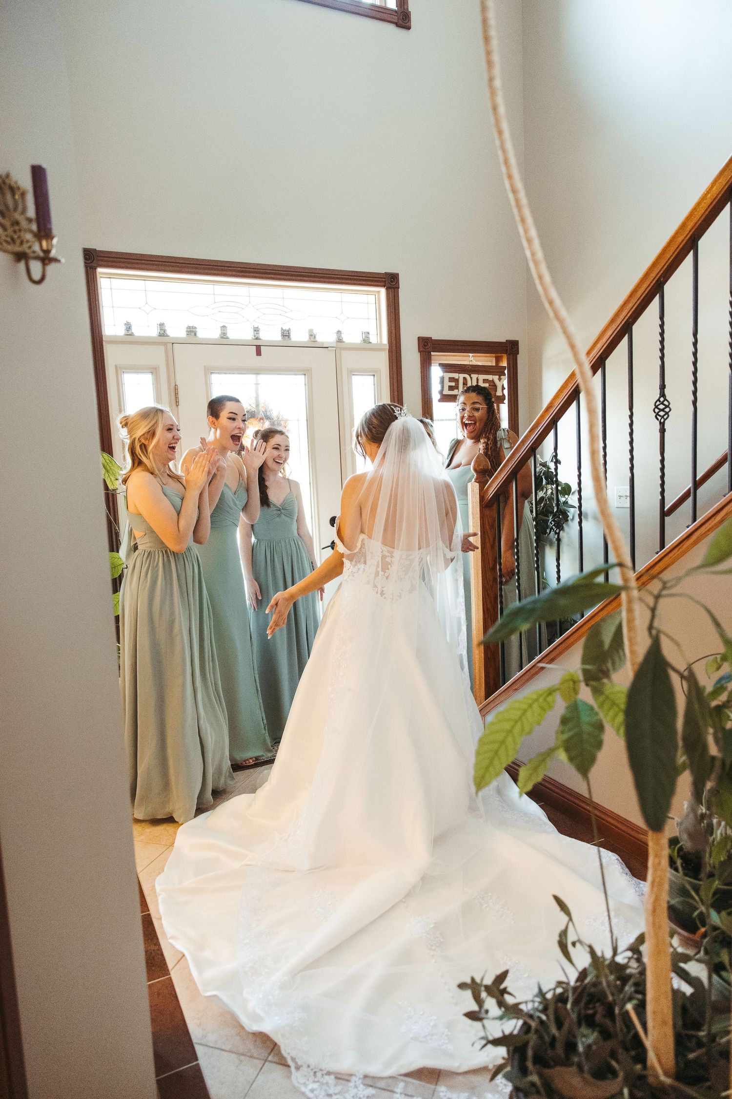 Bridesmaids in pastel dresses gather around bride in flowing white gown with veil on elegant staircase.