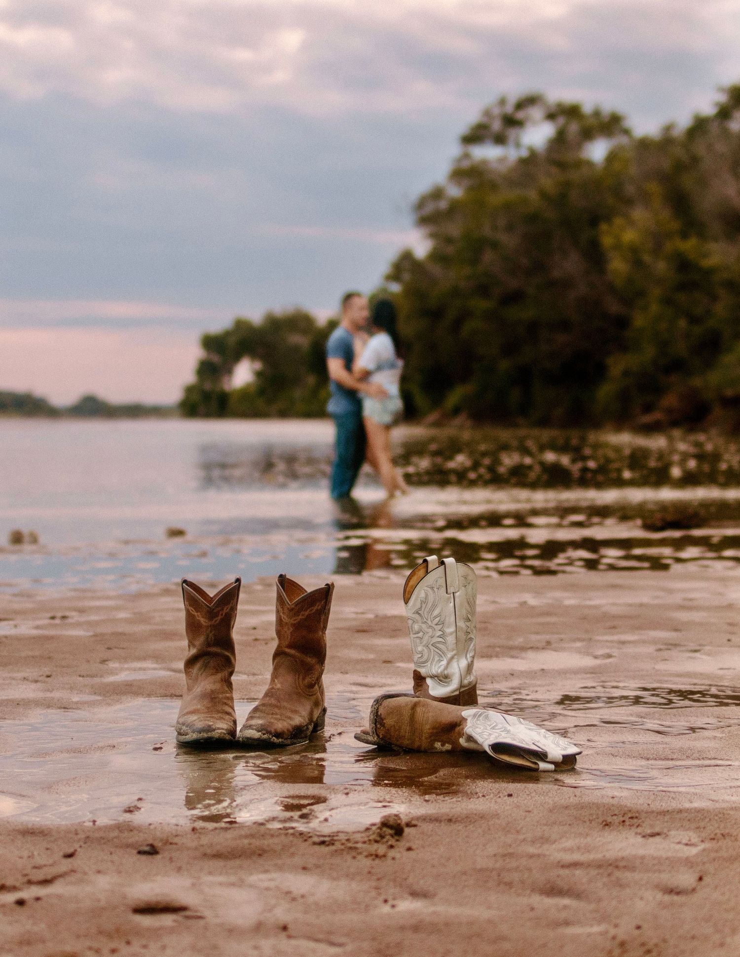 Three pairs of boots stand in a line on a sandy beach with water and trees visible in the background.