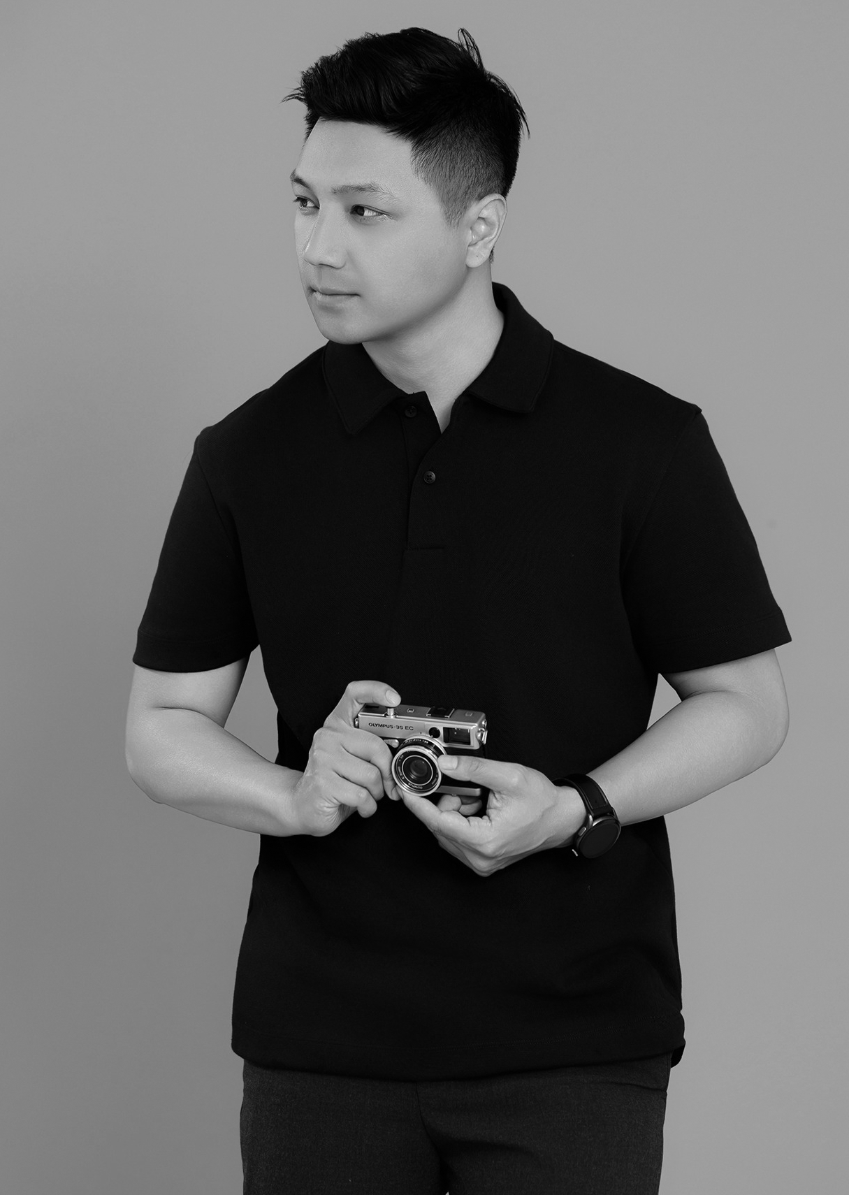 Black and white portrait of a person in a black polo shirt holding a vintage camera while looking to the side.
