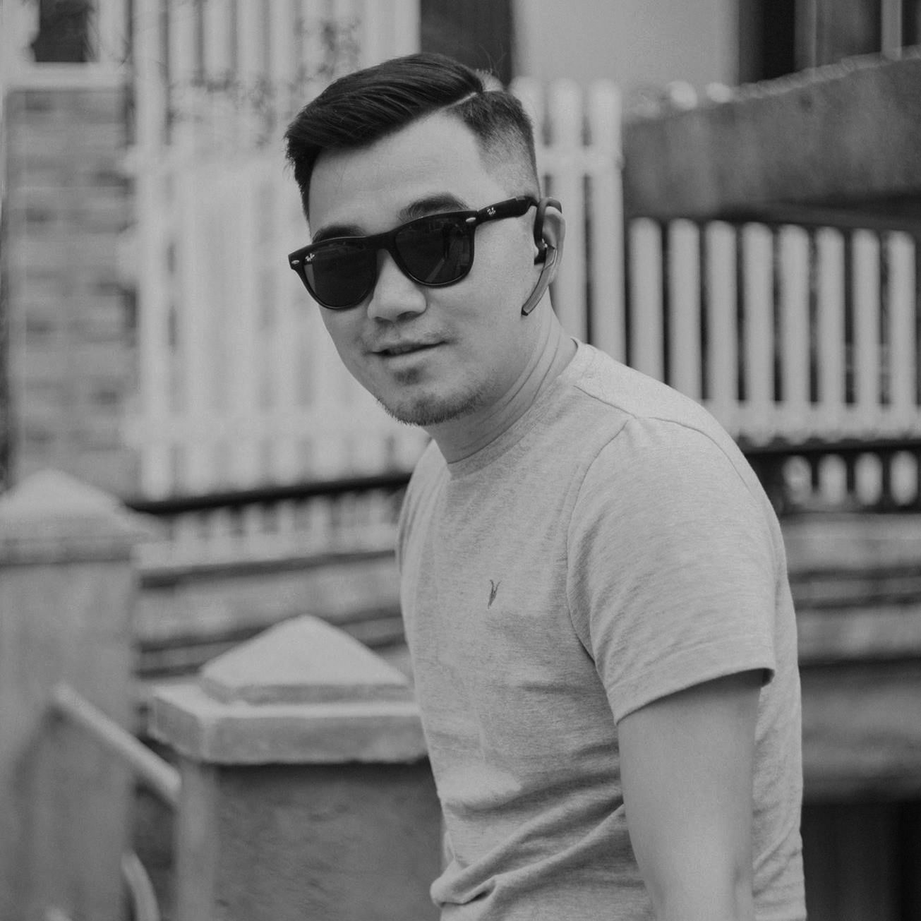 A black and white portrait of a person wearing sunglasses and a casual t-shirt in an urban setting.
