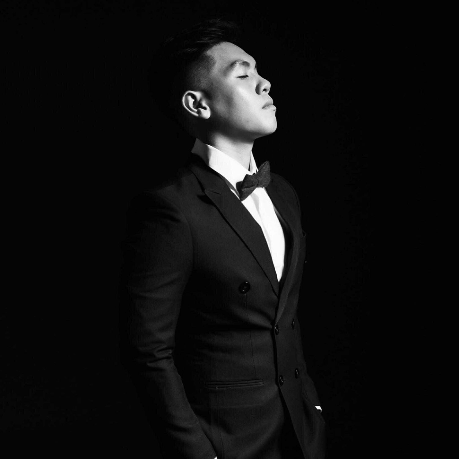 Dramatic black and white portrait of a figure wearing a tuxedo and bow tie with eyes closed against dark backdrop.