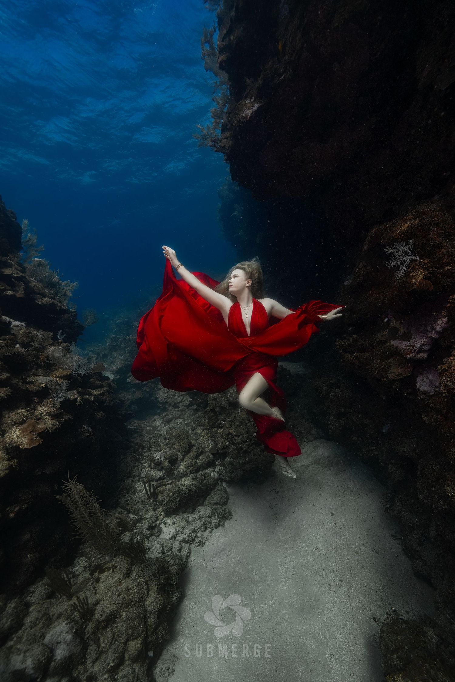 Underwater dancer in flowing red dress performs in deep blue ocean cave setting.