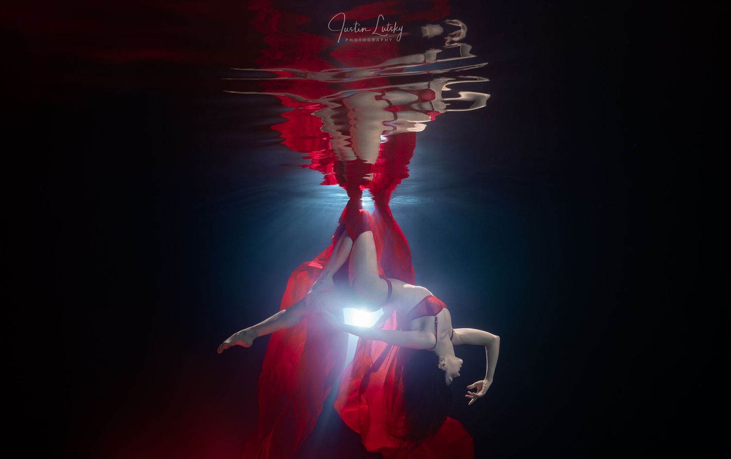 A figure in a flowing red dress appears underwater, illuminated by dramatic lighting and creating a reflection above.